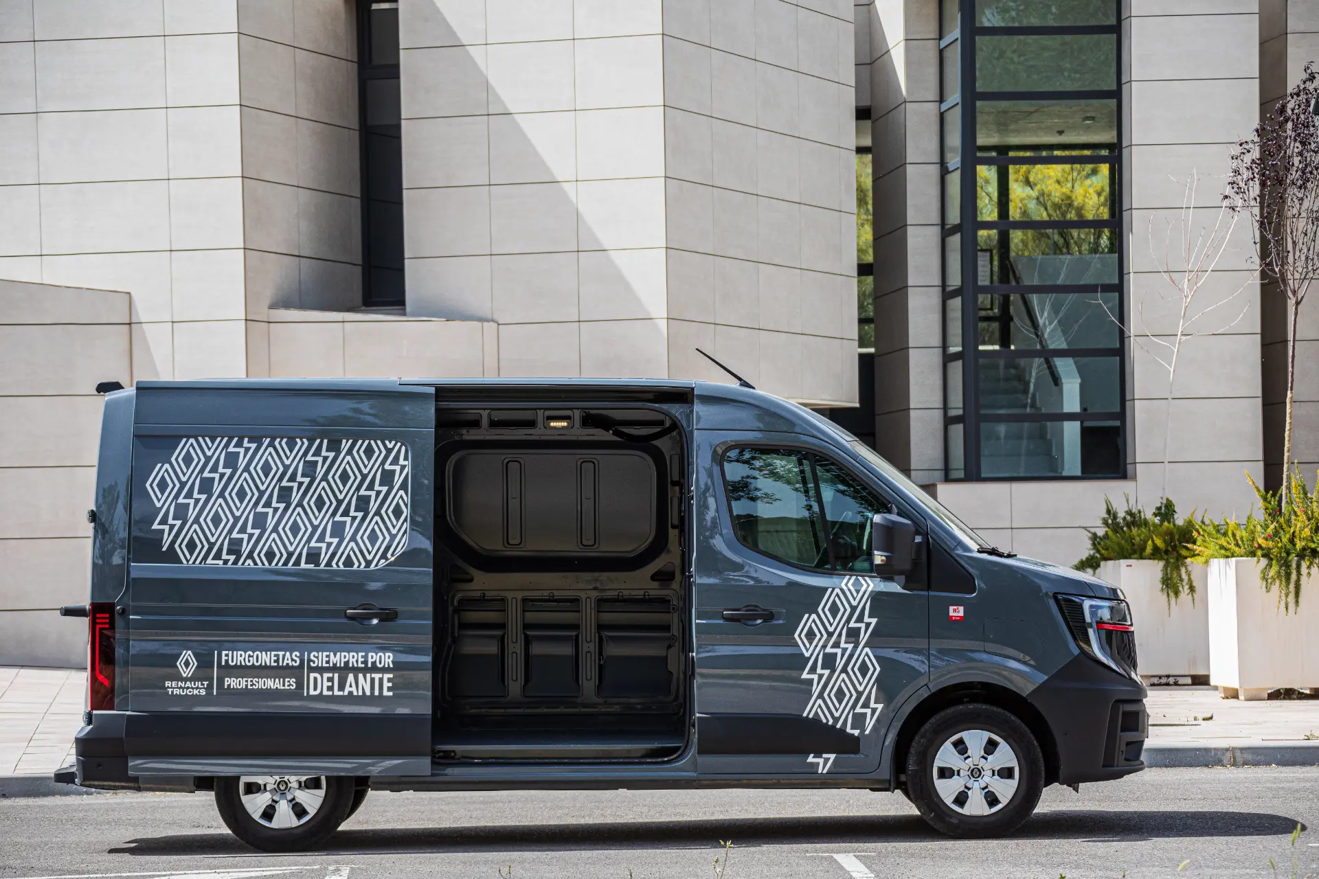 Vista lateral de un Renault Master furgón gris con una puerta corredera abierta, aparcado delante de un edificio moderno.