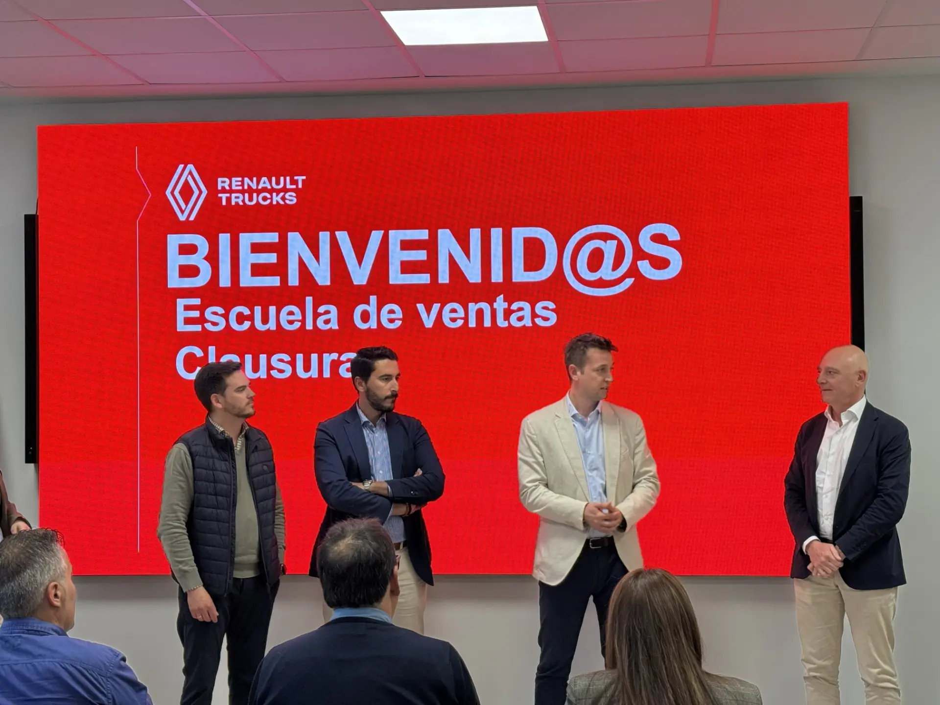Cuatro hombres de pie delante de un cartel rojo de Renault Trucks en un acto de clausura de una escuela de ventas, presentando a un público las ventajas de utilizar el borrador automático en la documentación de vehículos modernos.