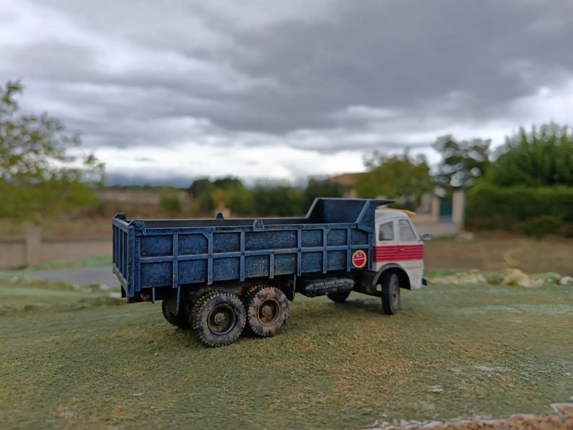Un camión volquete azul de juguete está sentado sobre la hierba con un cielo nublado y árboles de fondo, como si esperara su próxima carga de una obra de construcción de un borrador automático.