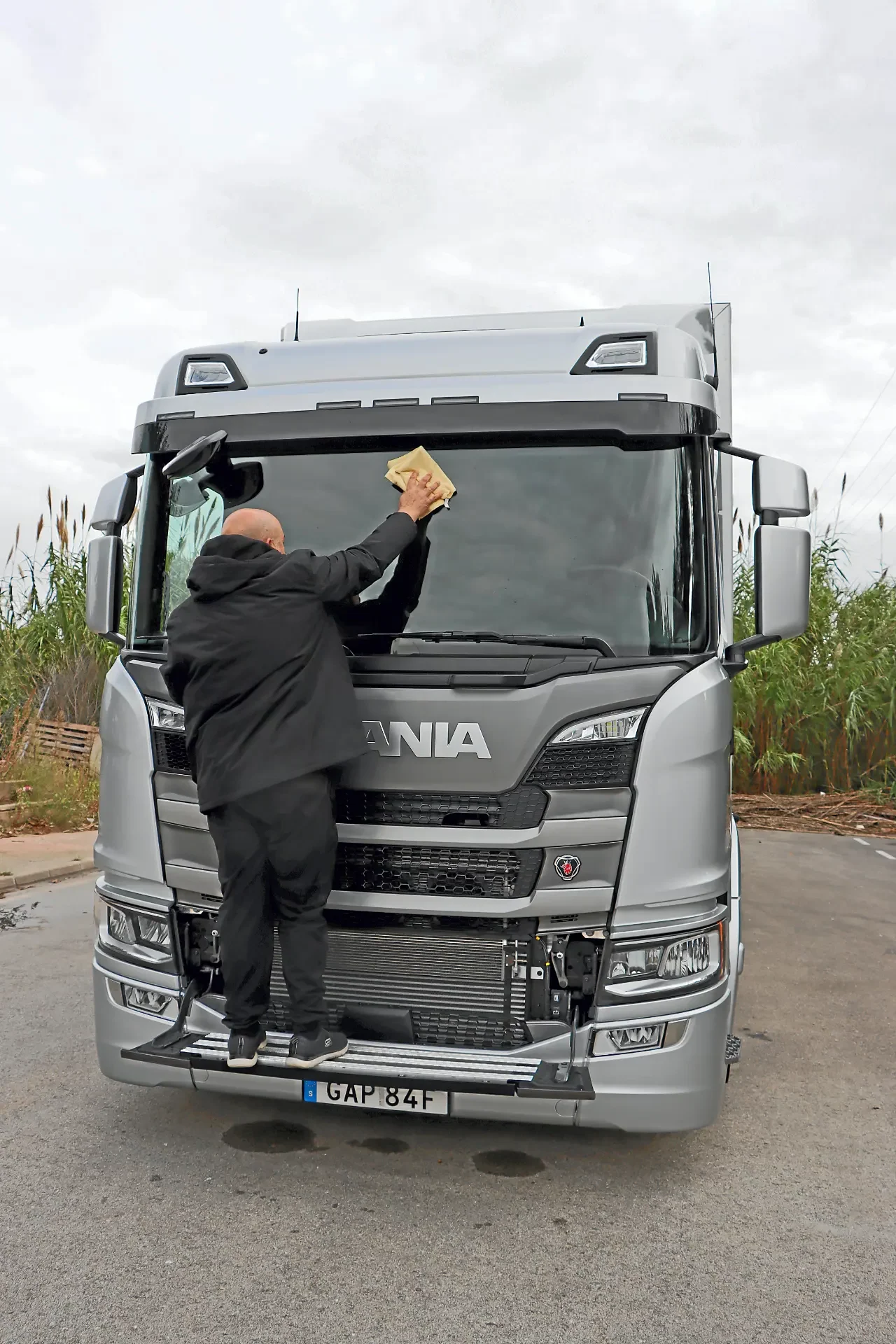 Un hombre trajeado limpia el parabrisas de un gran camión Scania 390G plateado aparcado al borde de la carretera, mostrando uno de los impresionantes camiones rígidos Scania.