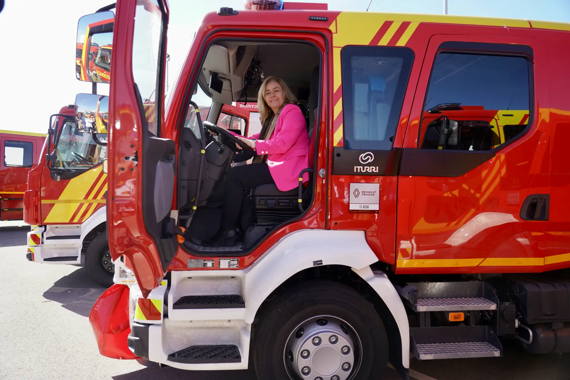 Una mujer vestida con una americana rosa se sienta en el asiento del conductor de un camión de bomberos rojo, con la puerta abierta como si estuviera lista para la acción, como un borrador automático a punto de entrar en acción.