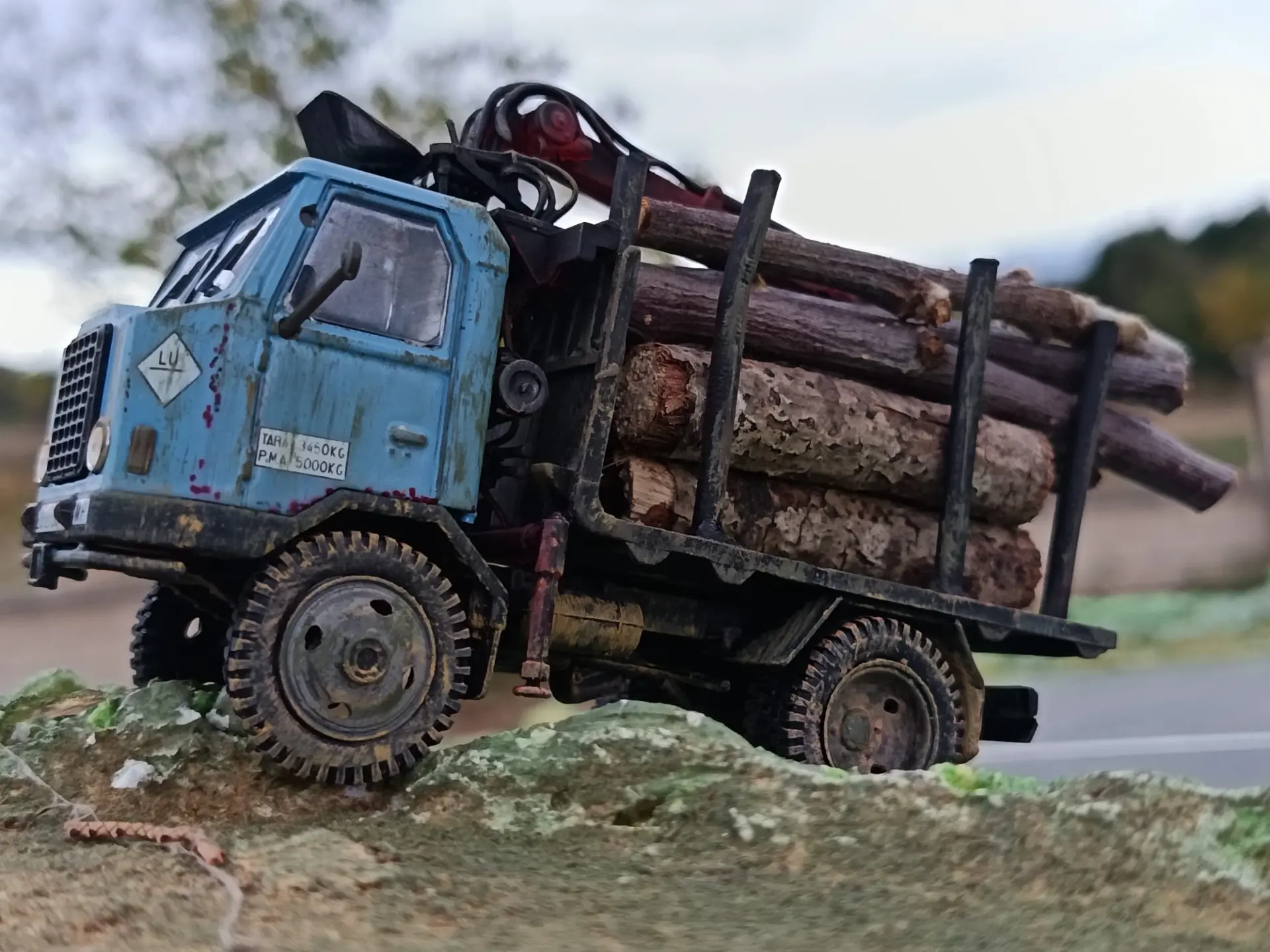 Un camión maderero de juguete azul, estilo carroceta escala 1:43, transporta troncos por terrenos abruptos al aire libre.