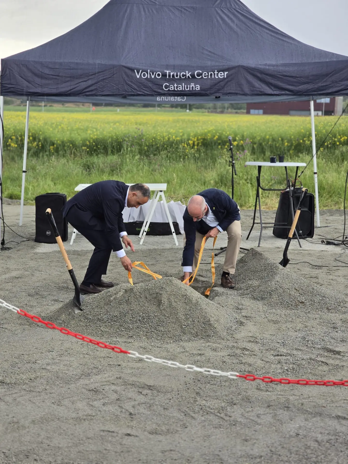 Dos hombres trajeados palean grava en un acto de colocación de la primera piedra bajo una carpa de Volvo Truck Center en Bescanó, celebrando el futuro de Truck Center Girona con Volvo Trucks.