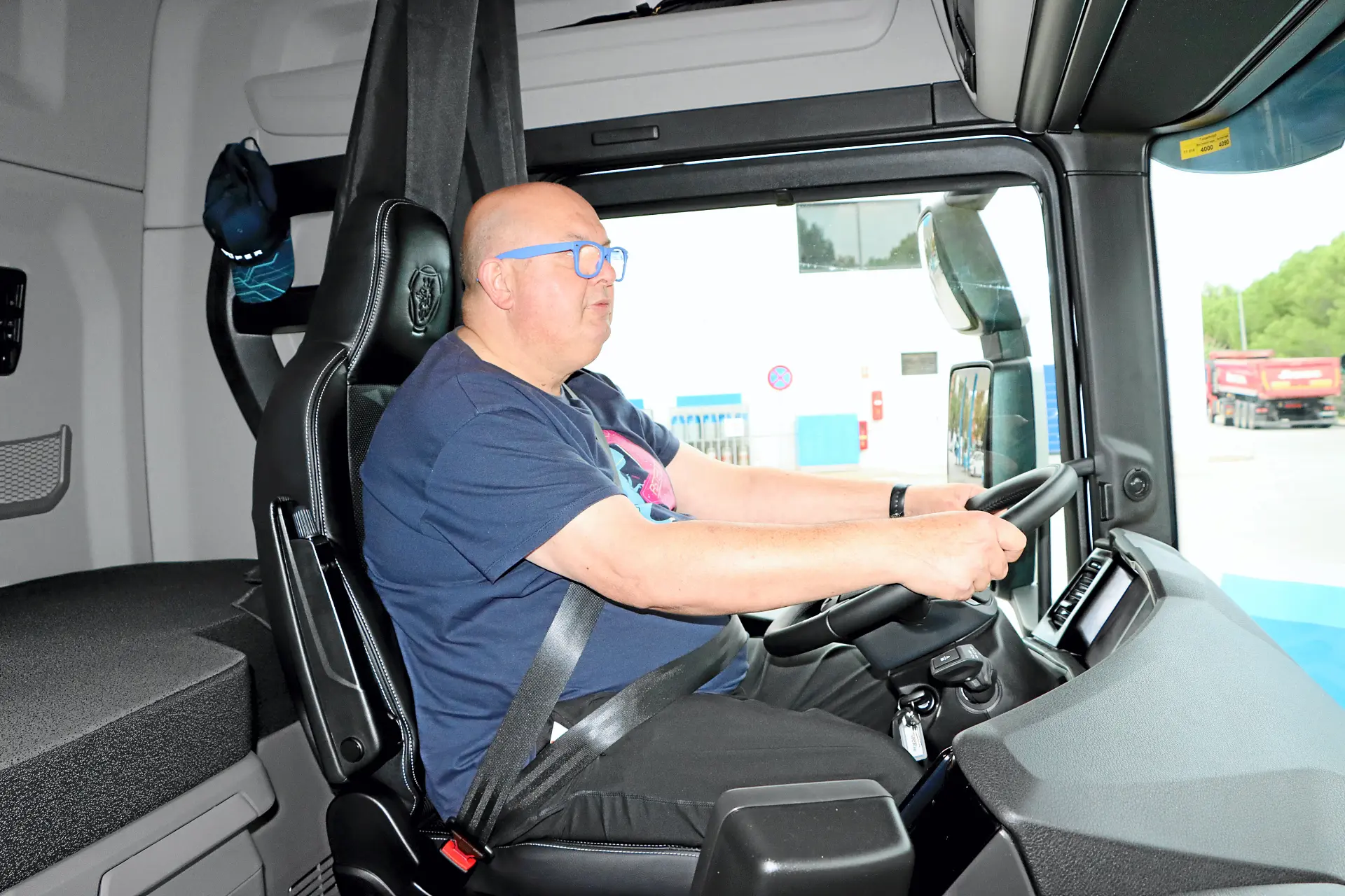 Un hombre con gafas conduce un camión, sentado en el asiento del conductor y con las manos en el volante, como si estuviera probando un borrador automático de navegación.