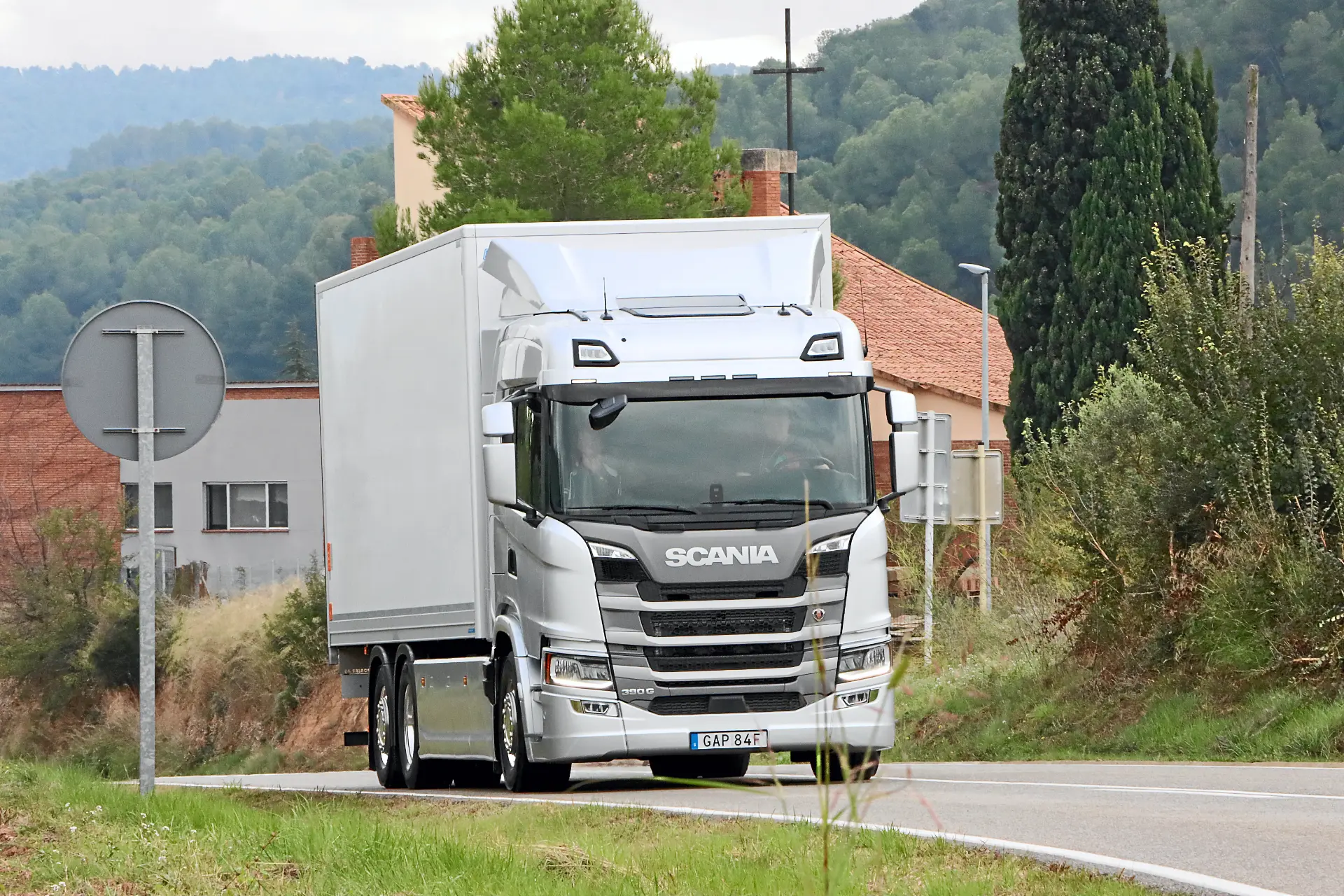 Un camión Scania blanco circula por una carretera rural con vegetación y edificios de fondo, con un sistema de borrador automático para mejorar la eficiencia.