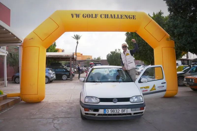 Un hombre atraviesa el techo solar de un VW Golf bajo un arco amarillo que reza "Volkswagen Golf Challenge", con el puño en alto en señal de triunfo tras completar 2.500 kilómetros de aventura 4x4.