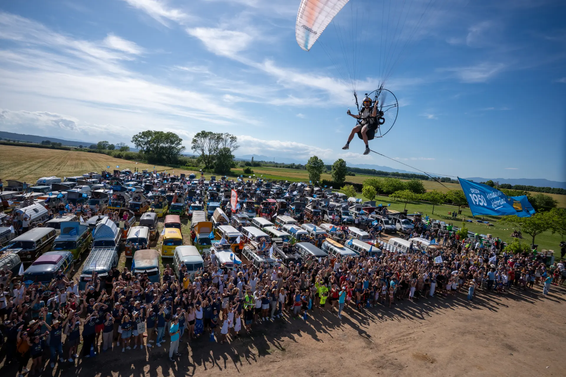 Un paramotor se eleva por encima de una animada multitud e hileras de Volkswagen Vehículos Comerciales en el evento FurgoVolkswagen 2026 en un día soleado.