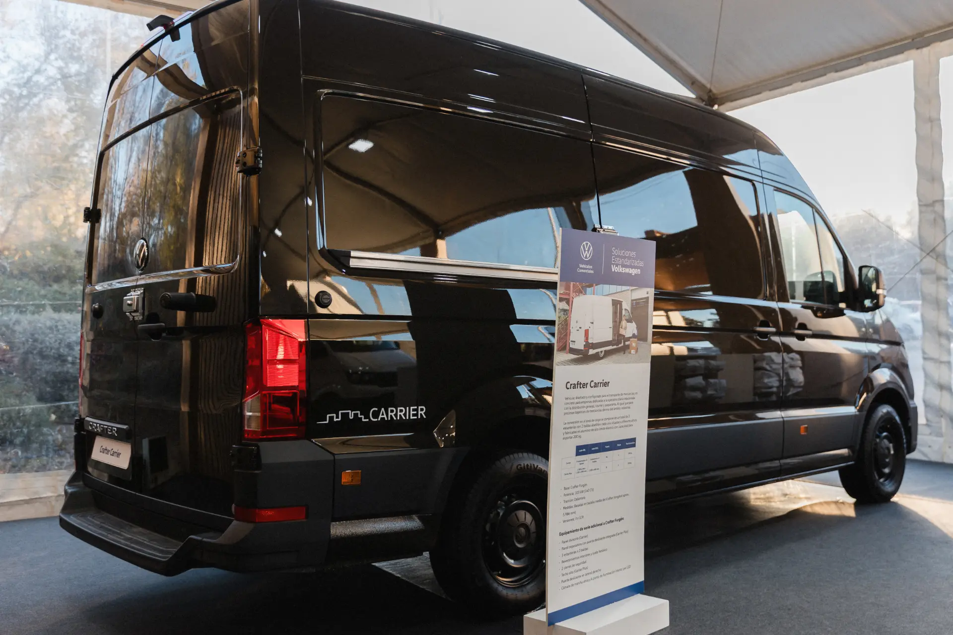Una furgoneta Volkswagen Carrier negra, de los Vehículos Comerciales, se exhibe en el interior junto a un cartel informativo bajo una carpa blanca, destacando las soluciones estandarizadas de Volkswagen.
