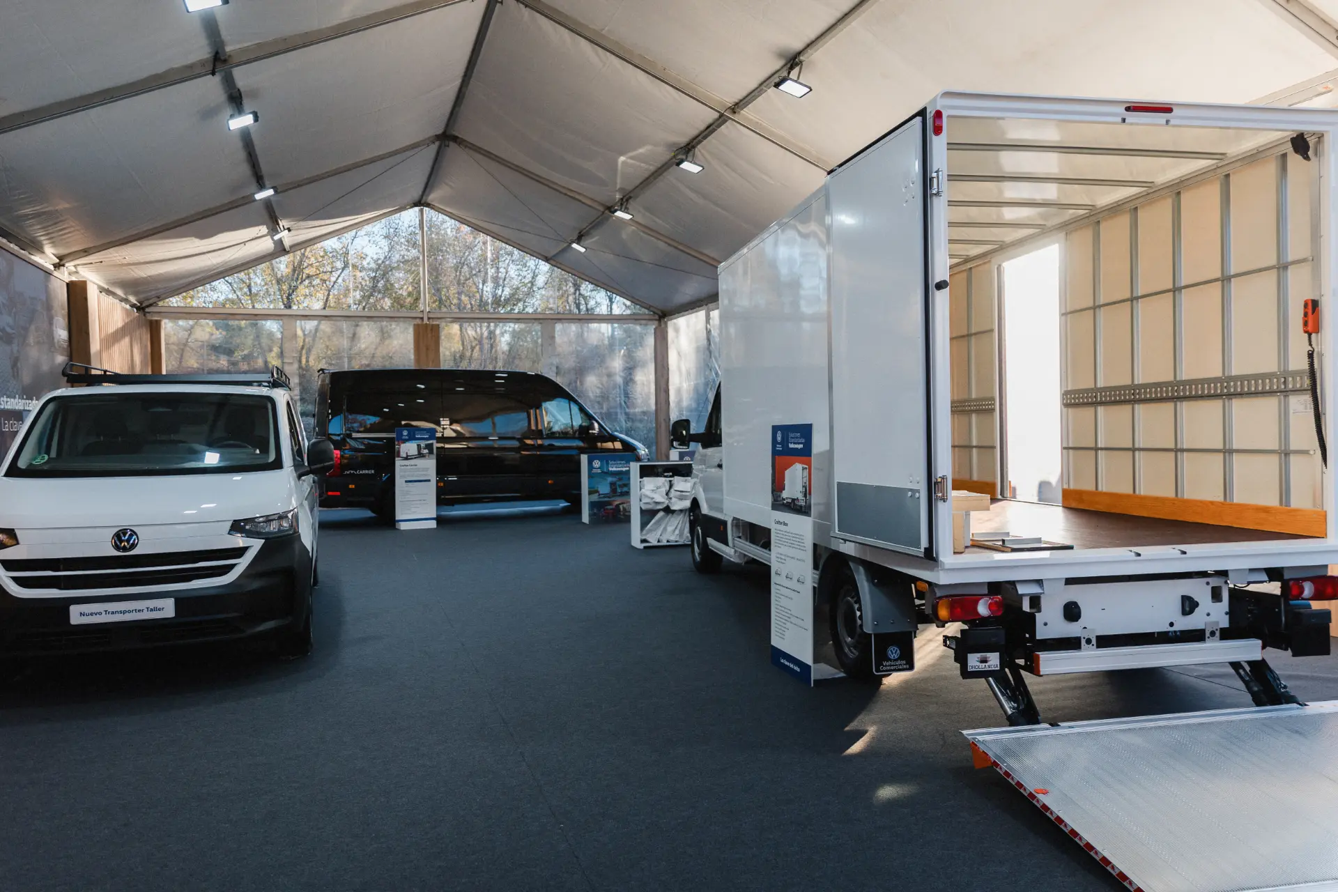 Tres Vehículos Comerciales, entre ellos una furgoneta Volkswagen y un camión caja, expuestos en el interior de una gran carpa showroom que muestra soluciones estandarizadas.