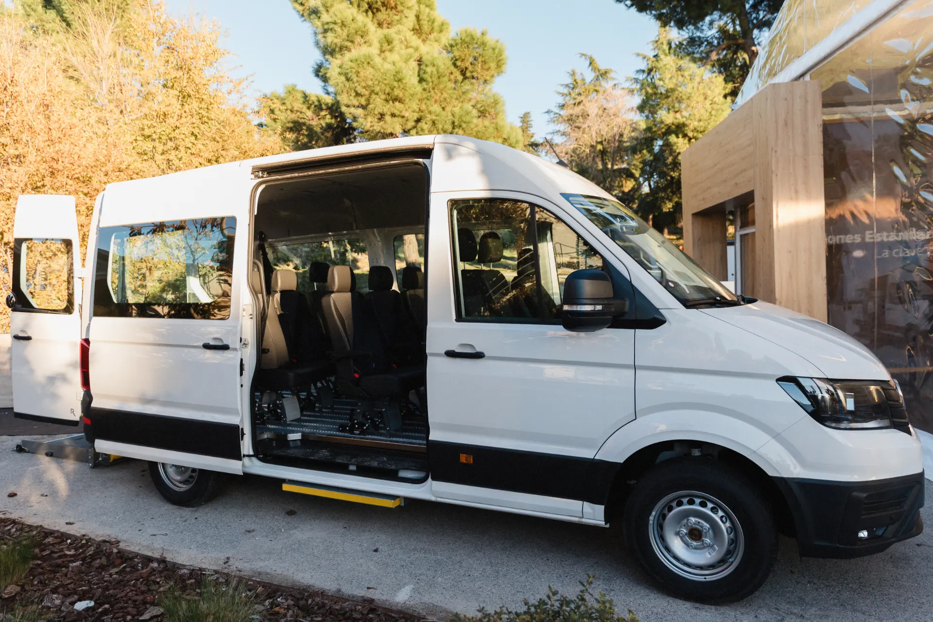 Furgoneta Volkswagen blanca de pasajeros con la puerta lateral abierta, dejando ver varias filas de asientos, aparcada al aire libre cerca de árboles y un edificio, ideal para quienes buscan soluciones estandarizadas en Vehículos Comerciales.