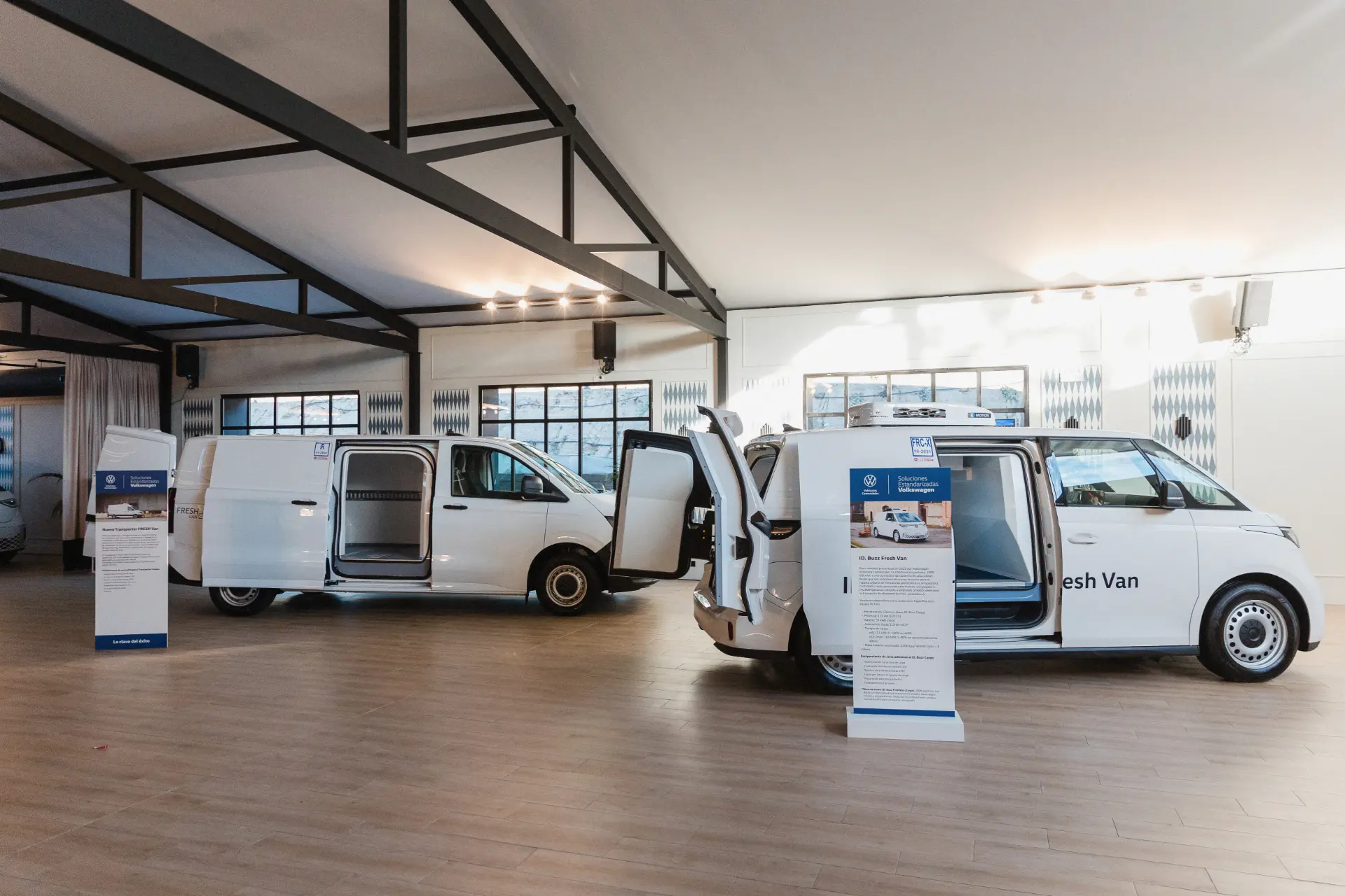 Dos furgonetas eléctricas blancas de Volkswagen Vehículos Comerciales se exhiben en el interior con carteles informativos delante de cada vehículo, mostrando soluciones estandarizadas para empresas modernas.