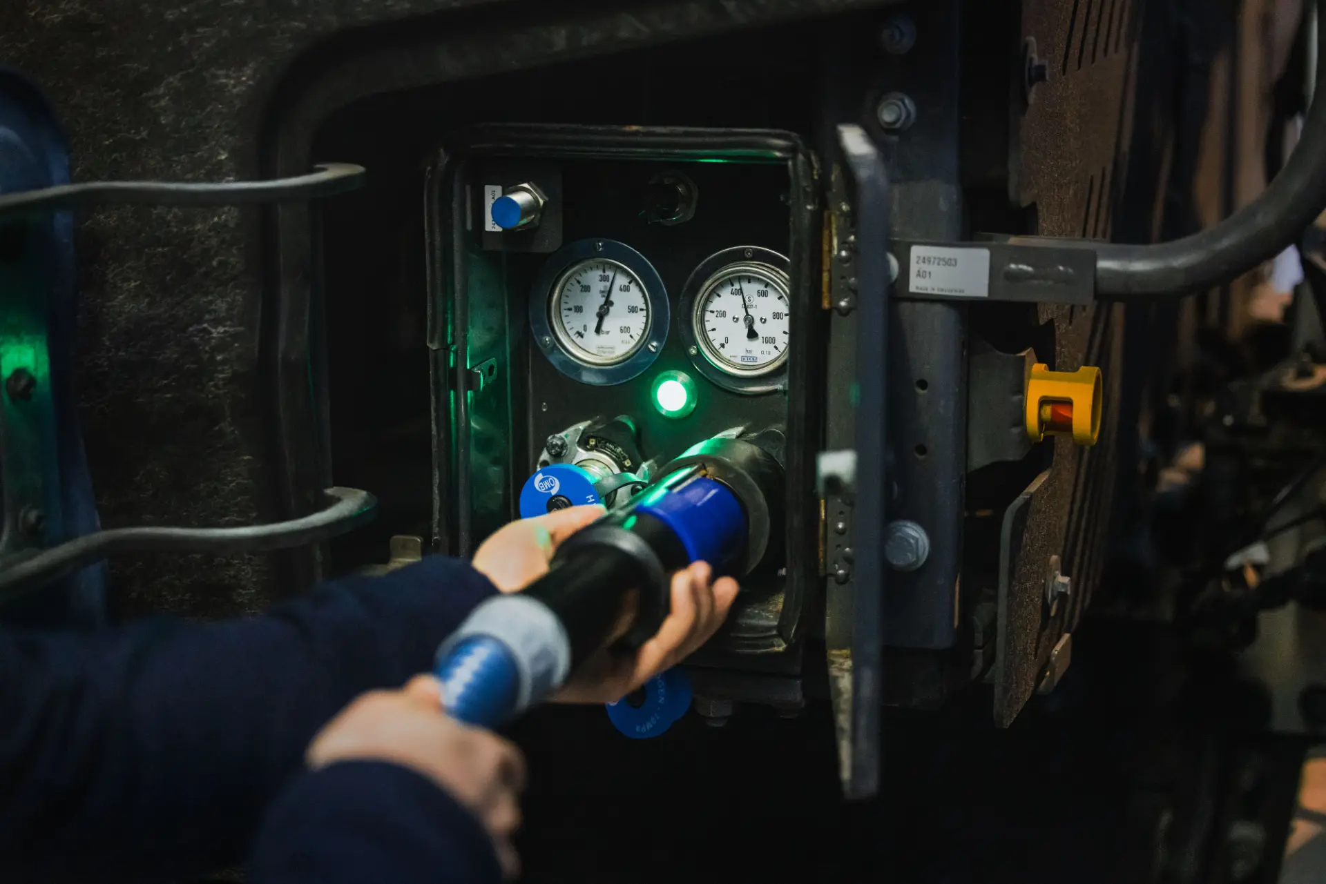 Manos conectando una boquilla de combustible a un puerto con manómetros y luces indicadoras visibles, ilustrando el proceso de repostaje para camiones Volvo propulsados por hidrógeno.