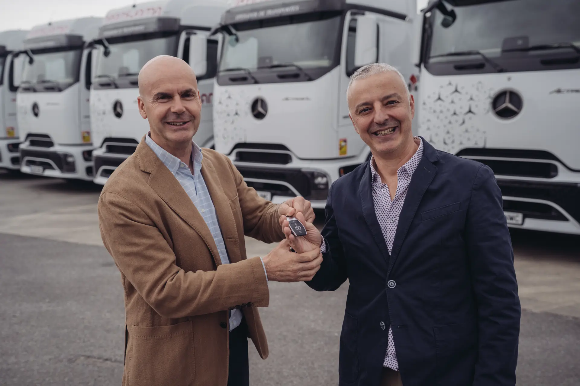 Dos hombres sonríen mientras sostienen una llave de Mercedes-Benz delante de varios camiones blancos, mostrando la nueva flota Mercedes-Benz Actros 1853 LS StreamSpace ProCabin de O.T. Transjama, conocida por su eficiencia energética.