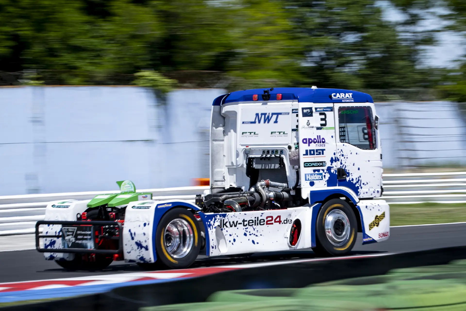 Un camión de carreras blanco y azul patrocinado por DT Spare Parts acelera en un circuito de carreras de camiones, con Sascha Lenz al volante y los logotipos de los patrocinadores visibles sobre un fondo borroso.
