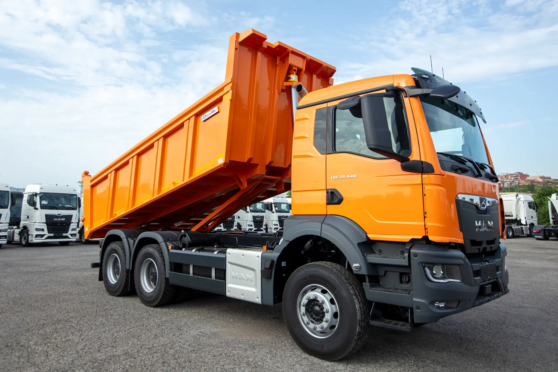 Camión volquete MAN de color naranja brillante con la caja levantada, aparcado sobre el asfalto bajo un cielo parcialmente nublado: un ejemplo de camiones de obra de calidad fabricados por expertos carroceros.