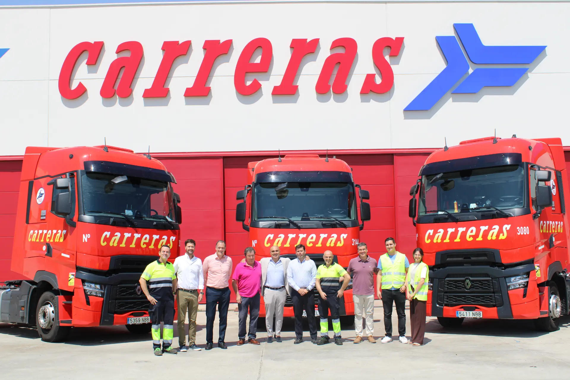 Nueve personas de pie delante de dos camiones rojos con el logotipo de "carreras" en un edificio detrás de ellos, como si estuvieran listas para una foto de equipo de borrador automático.