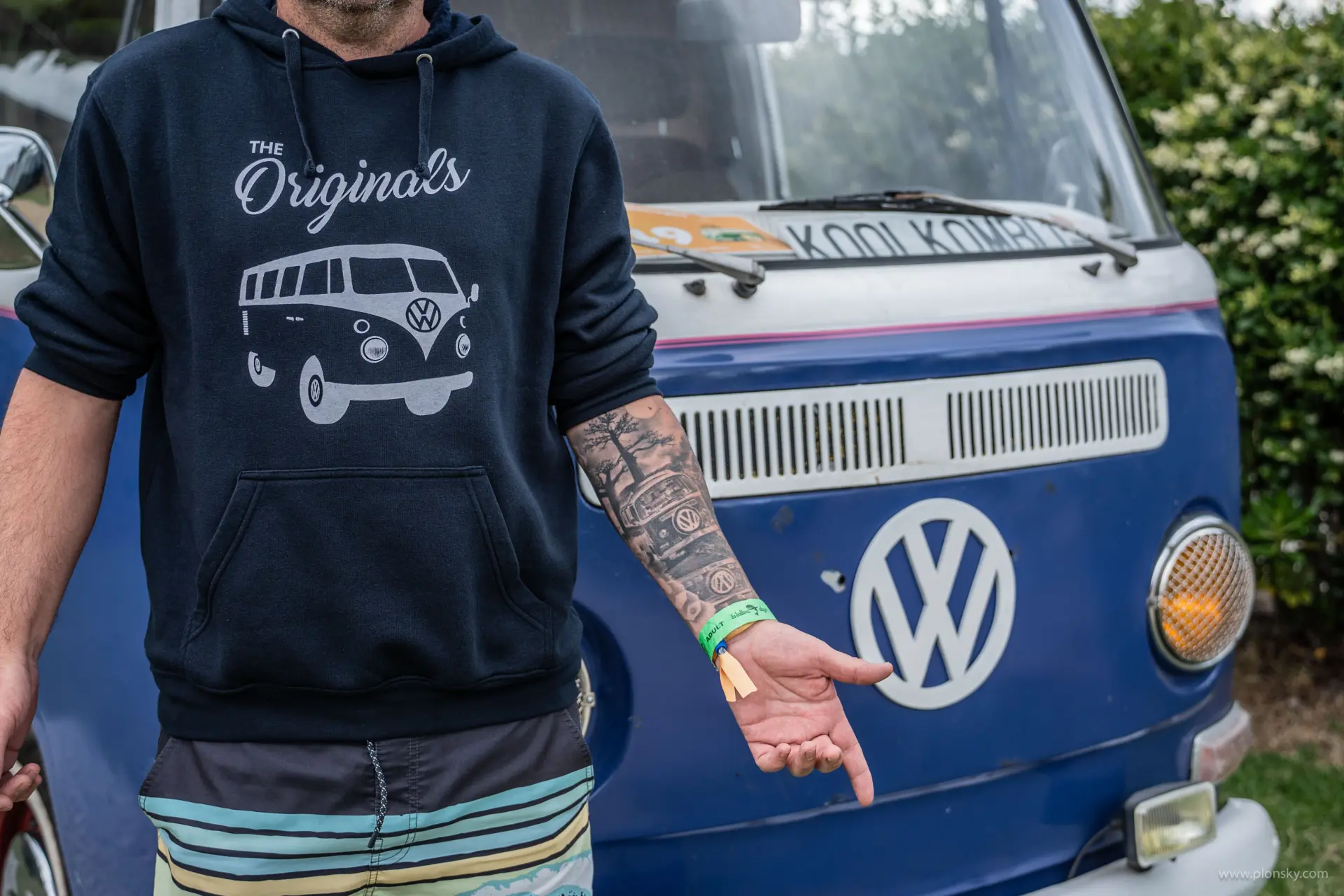 Una persona con una sudadera con capucha VW junto a una furgoneta clásica azul FurgoVolkswagen, con sus detallados tatuajes en los brazos a la vista: un auténtico fan de las furgos vintage y del estilo Volkswagen.