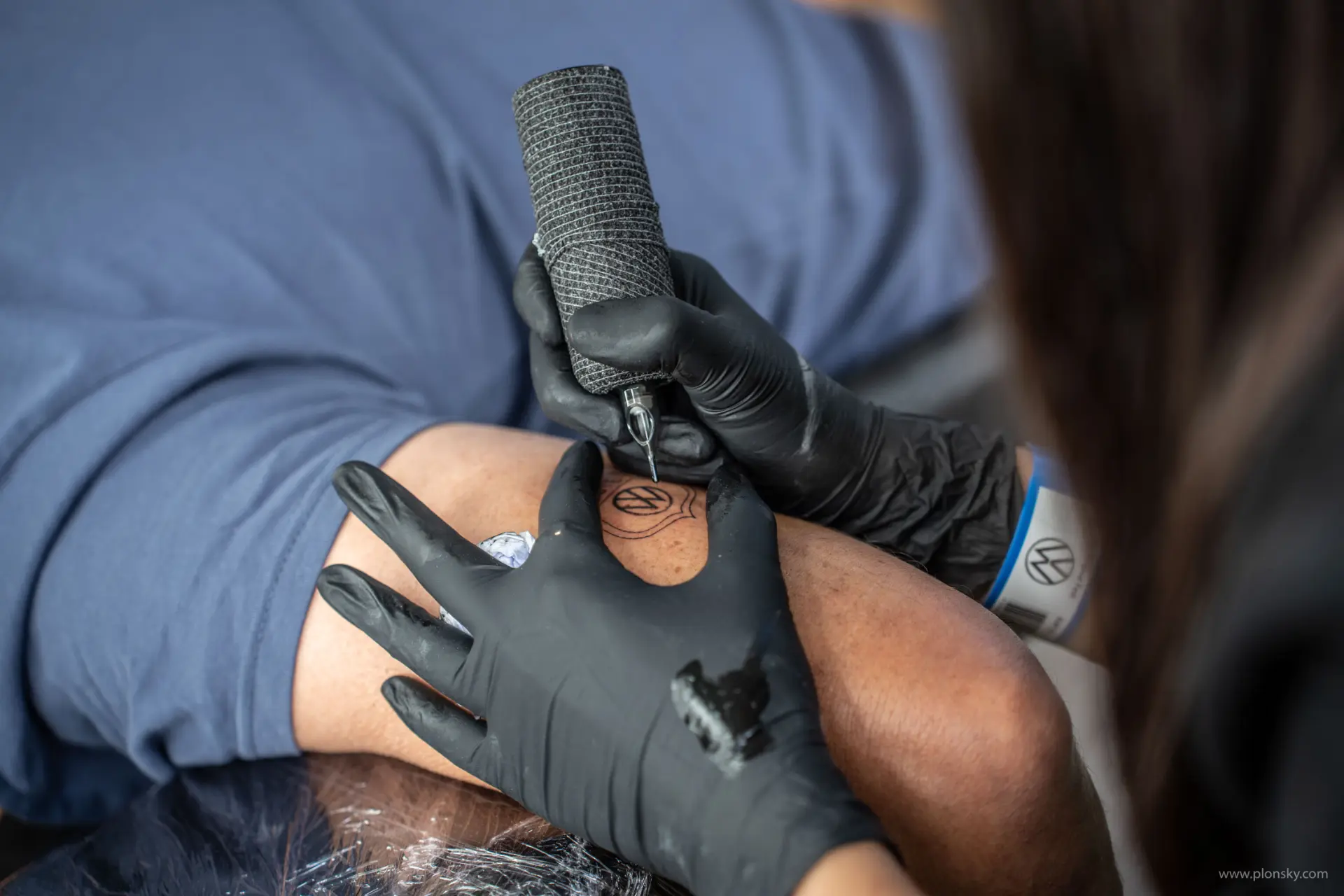 Artista del tatuaje con guantes negros tatuando un diseño circular Bulli, inspirado en el clásico FurgoVolkswagen, en el brazo de una persona.