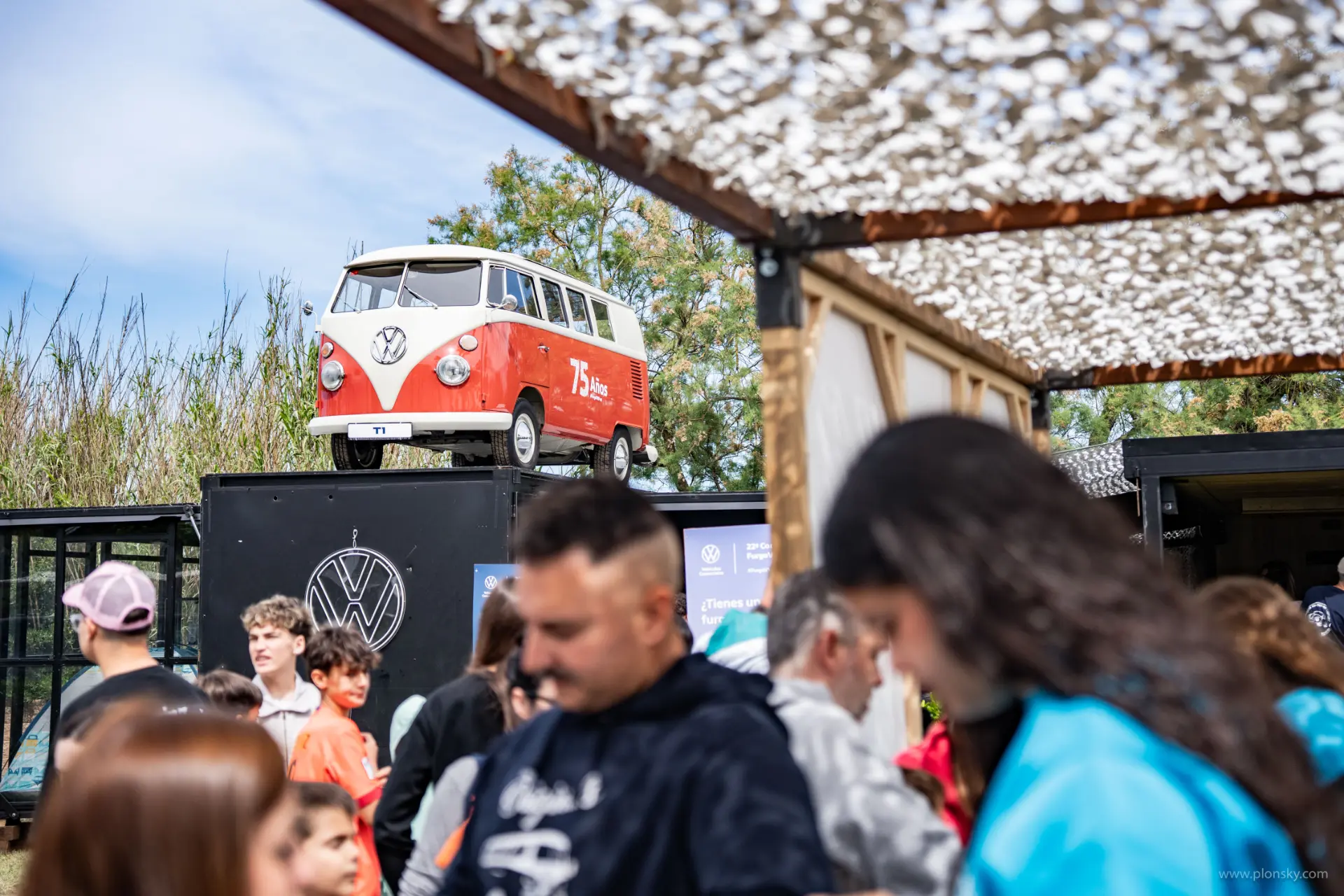 La gente se reúne en un evento al aire libre mientras un modelo clásico de furgoneta Volkswagen FurgoVolkswagen se exhibe en una plataforma por encima de la multitud.