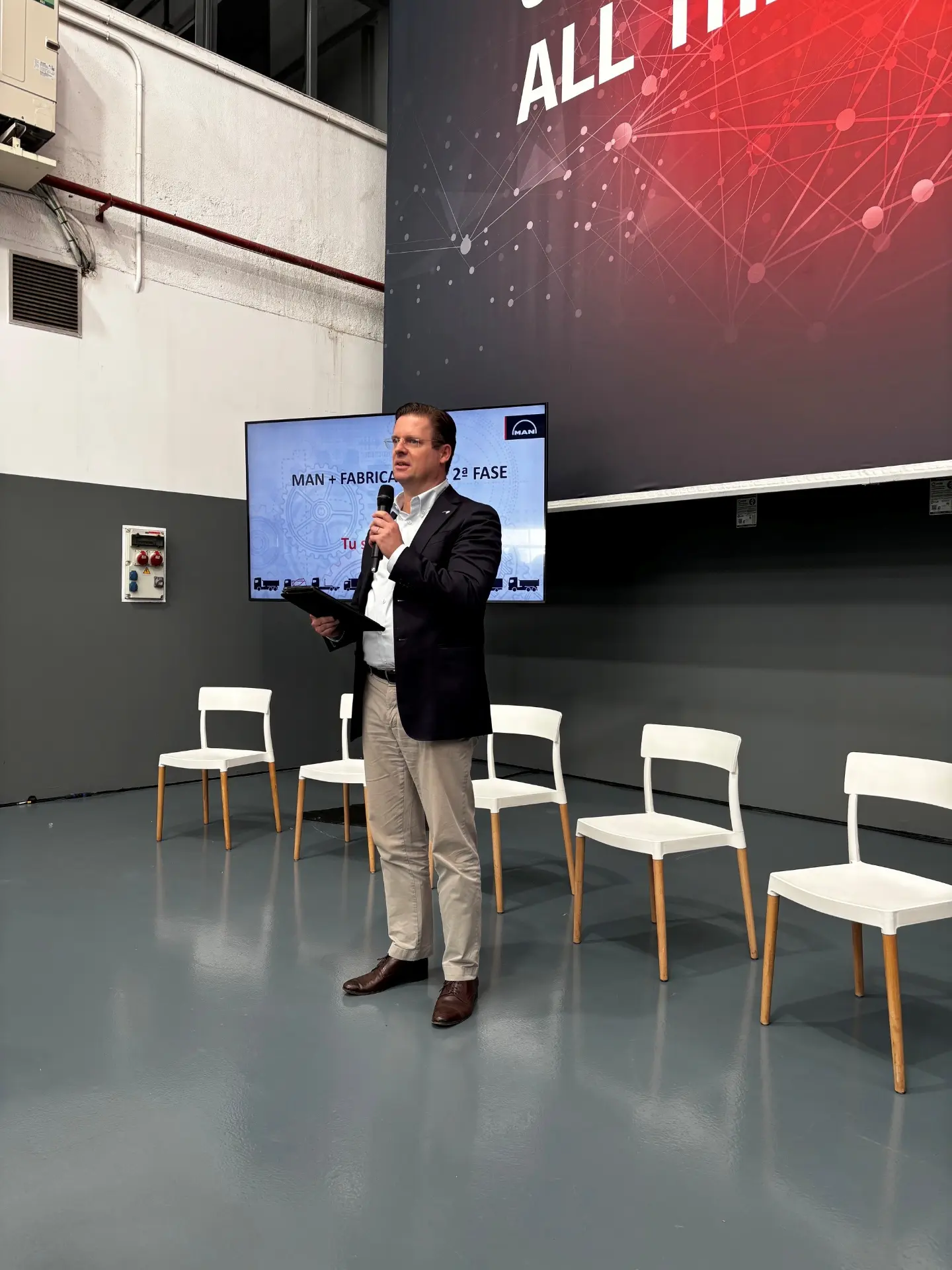 Un hombre trajeado habla en el escenario sobre MAN vehículos rígidos, con cuatro sillas blancas y una pantalla de presentación detrás de él.