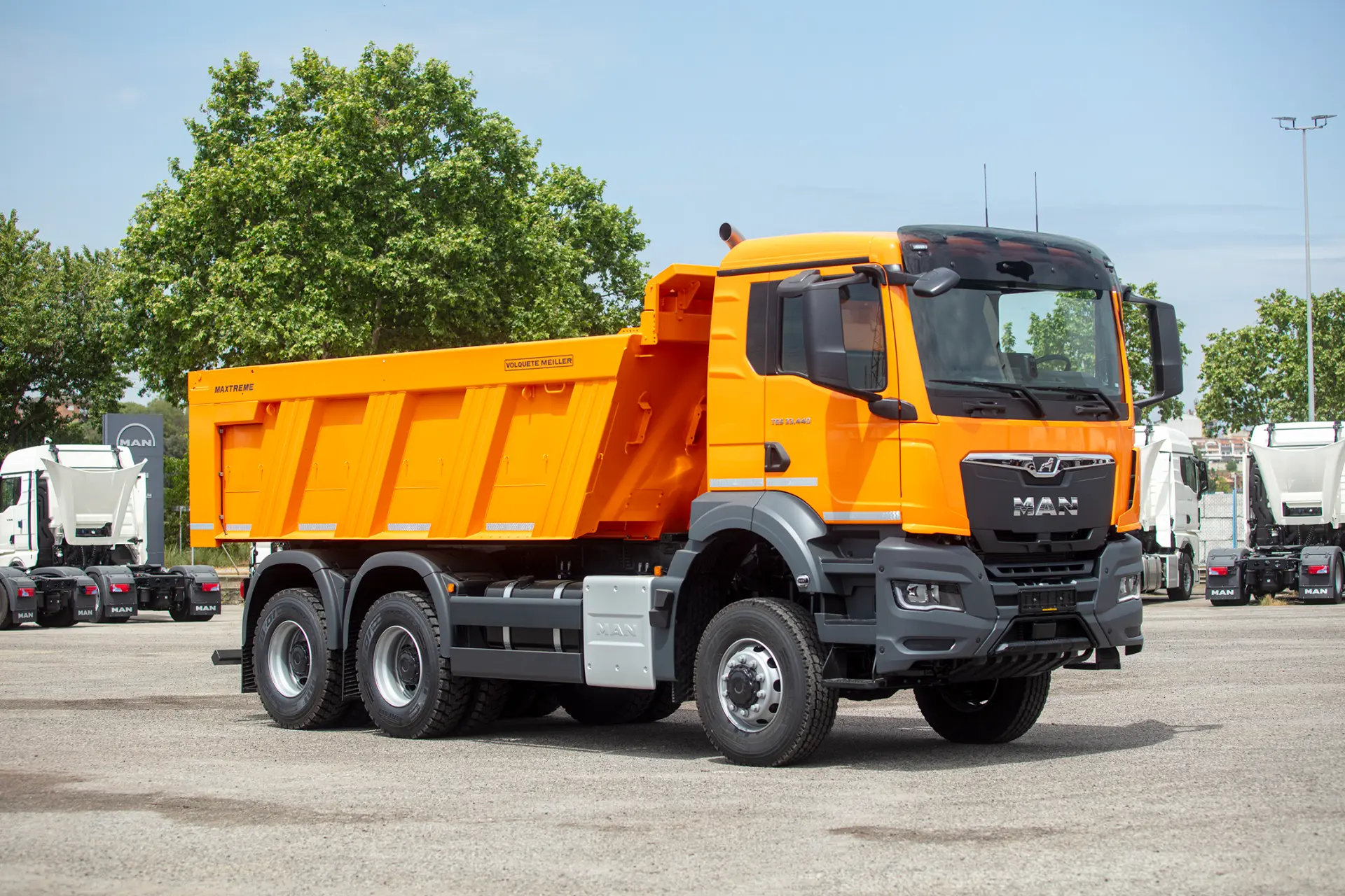 Volquete MAN amarillo brillante por carroceros estacionado en un terreno pavimentado, con árboles y otros vehículos rígidos de MAN Truck & Bus Iberia en el fondo.