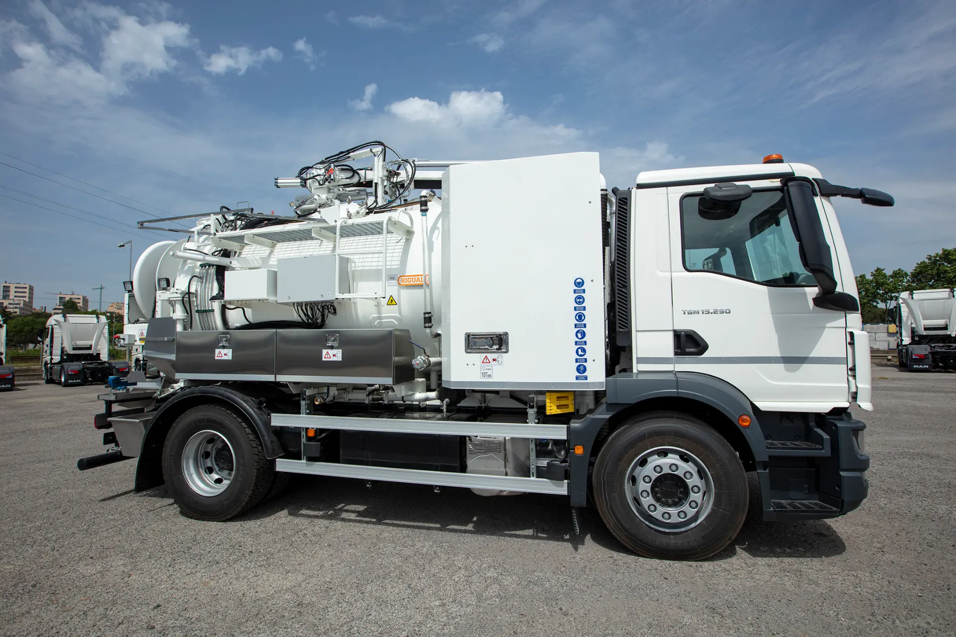 Un gran camión MAN blanco de aspiración industrial de carroceros, diseñado para camiones de obra, está aparcado en un solar asfaltado bajo un cielo parcialmente nublado.