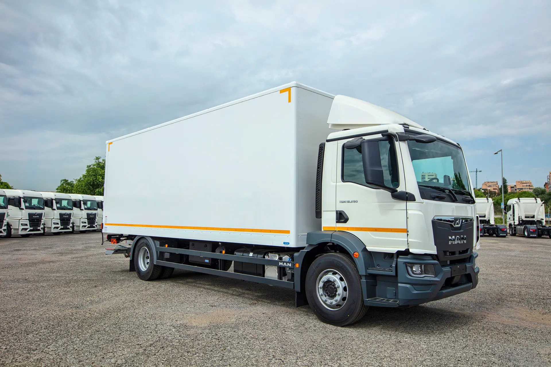 Un camión MAN blanco aparcado sobre el asfalto con más camiones visibles al fondo bajo un cielo nublado, mostrando la solución 360 de MAN Truck & Bus Iberia para las necesidades de transporte modernas.