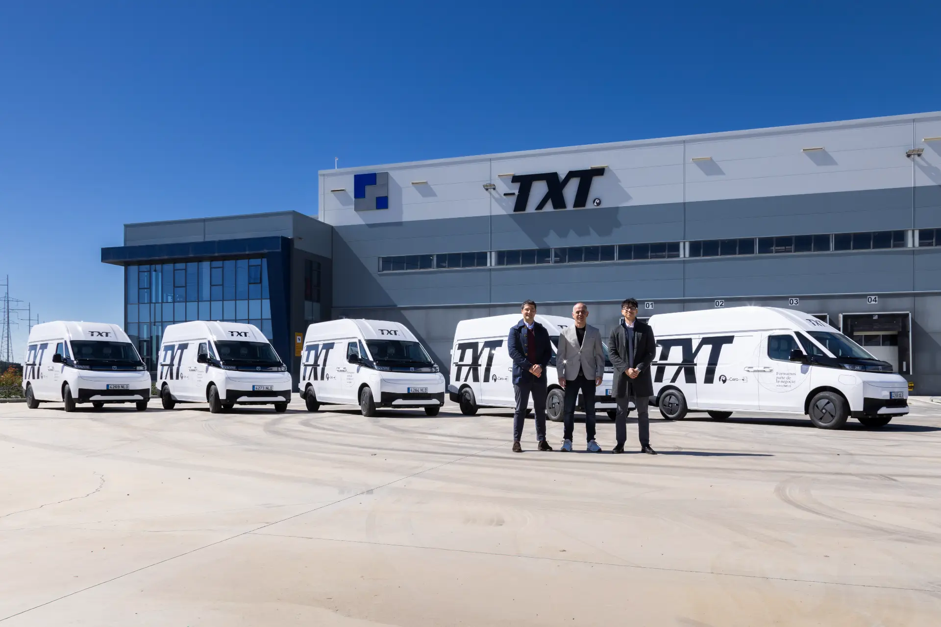 Dos hombres trajeados delante de cinco furgonetas TXT blancas, con el logotipo "Borrador automático", aparcadas frente a una moderna nave industrial.