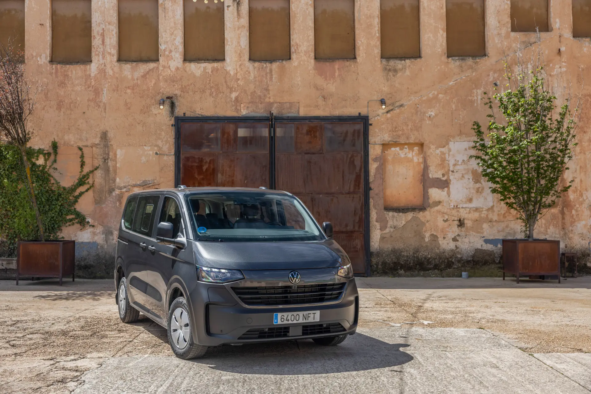 Una furgoneta gris de Volkswagen Vehículos Comerciales está aparcada frente a un desgastado edificio beige con ventanas enrejadas y un gran portón de madera, mostrando la creciente presencia de la marca en el mercado electrificado.