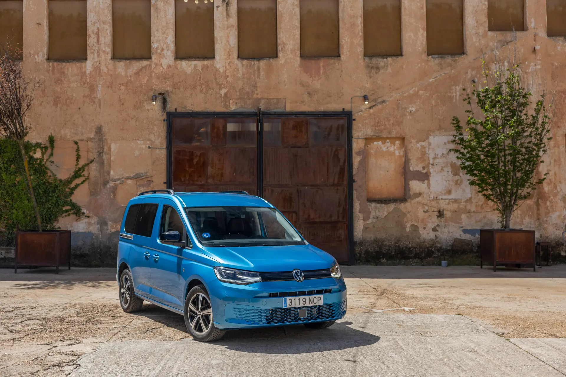 Una furgoneta Volkswagen Vehículos Comerciales azul está aparcada delante de un viejo edificio beige con ventanas y plantas junto a las paredes.