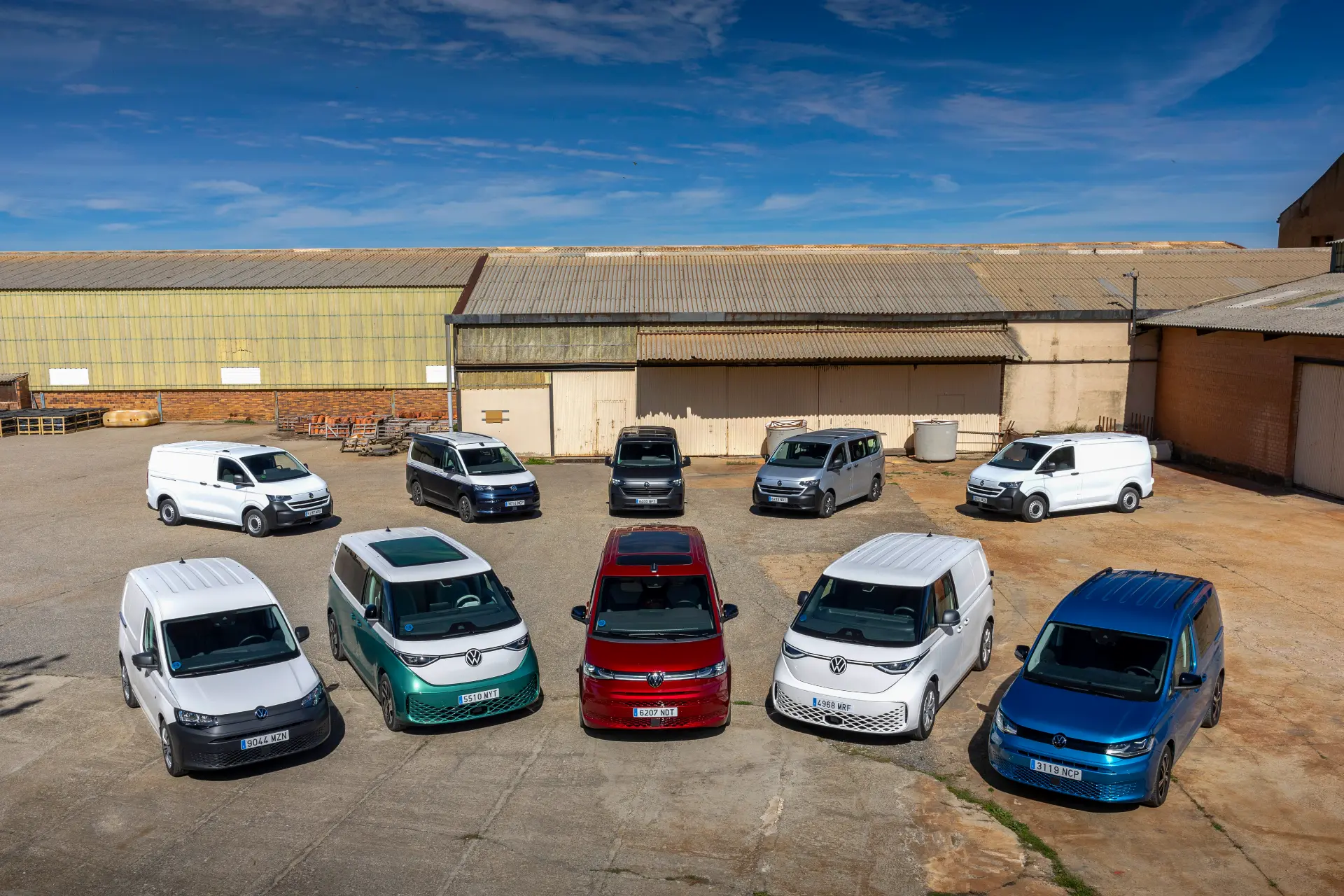 Nueve furgonetas de Volkswagen Vehículos Comerciales de diferentes colores están aparcadas en semicírculo frente a naves industriales bajo un cielo azul, mostrando el compromiso de la marca de electrificar su mercado con una diversa gama electrificada.