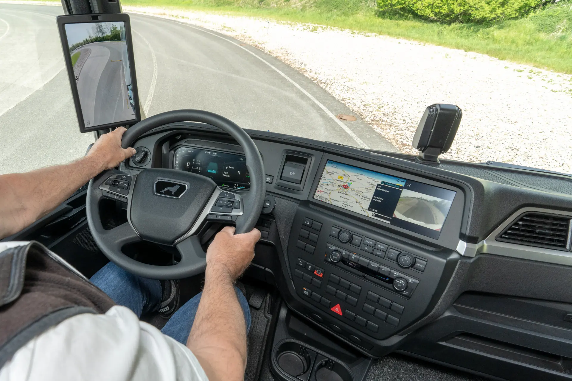 Una persona conduce un camión con un salpicadero digital con Borrador automático, pantallas que muestran mapas y una cámara de visión trasera, todo ello mientras navega por una carretera curva.