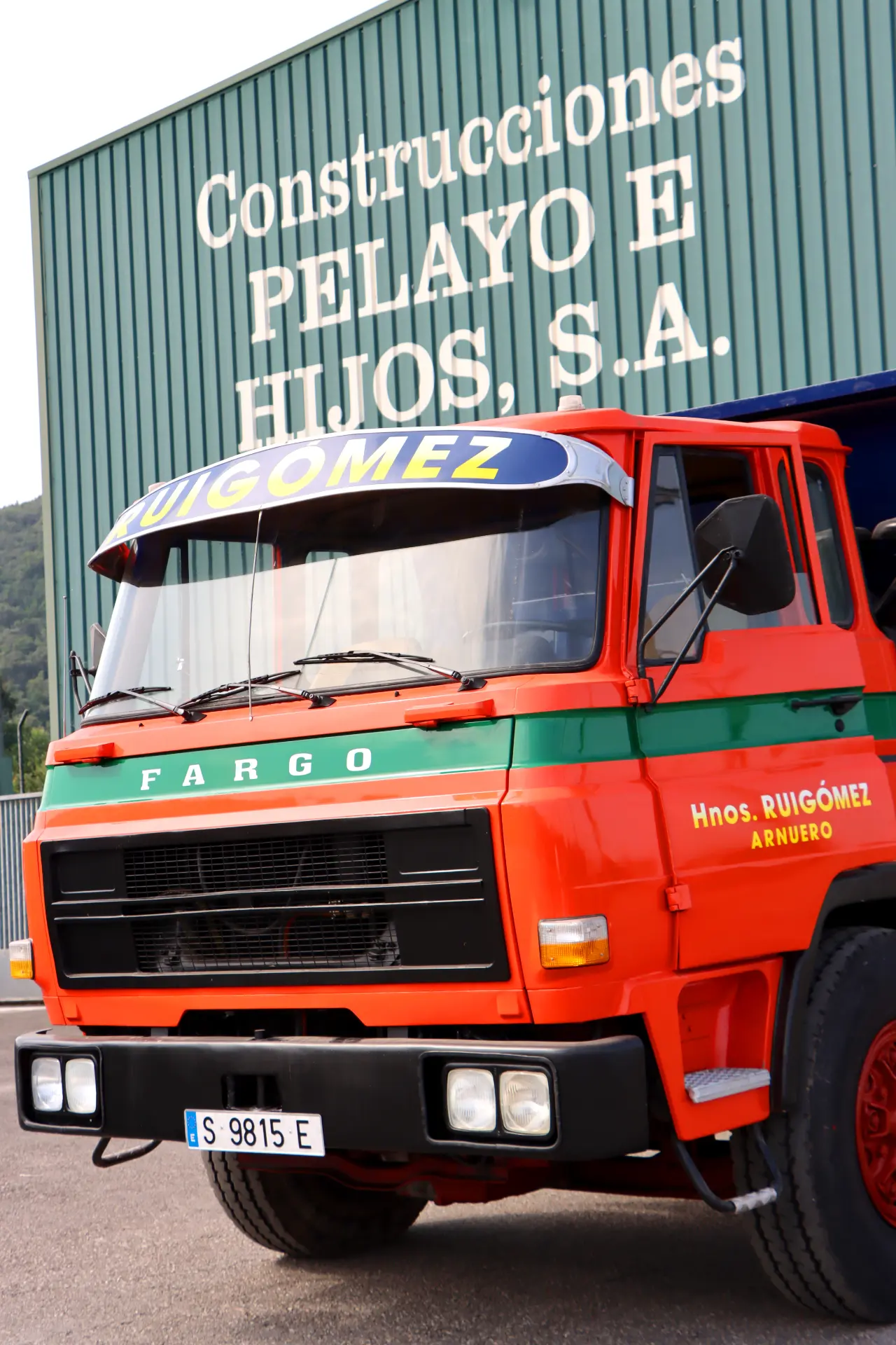 Camión Fargo rojo y verde, un gran ejemplo de camiones clásicos, aparcado delante de un edificio verde con el letrero "Construcciones PELAYO E HIJOS, S.A.".