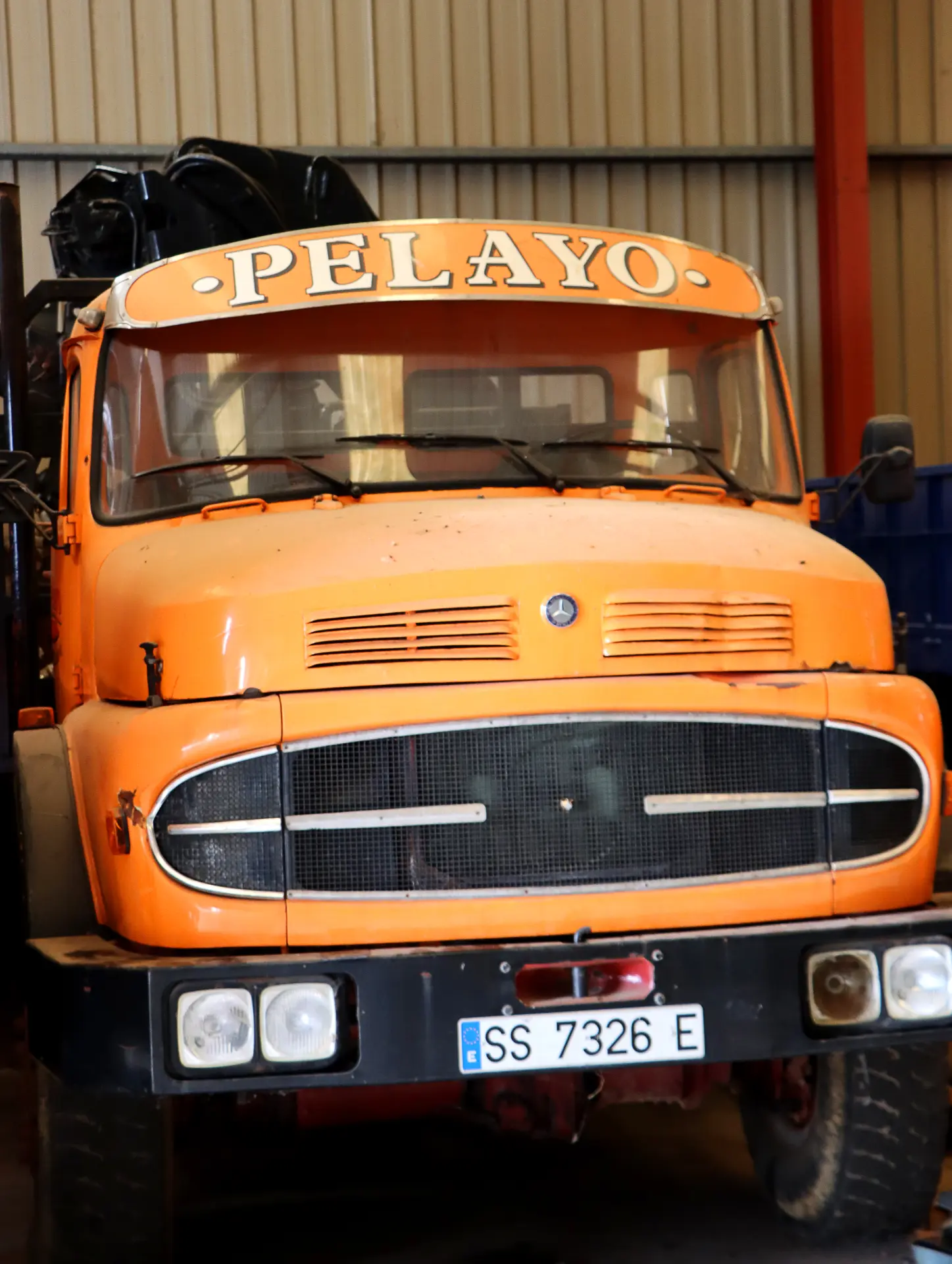 Camión Barreiros naranja de época con "PELAYO" en el parabrisas, aparcado en el interior y con la matrícula delantera visible. Un ejemplo clásico de camiones Barreiros.