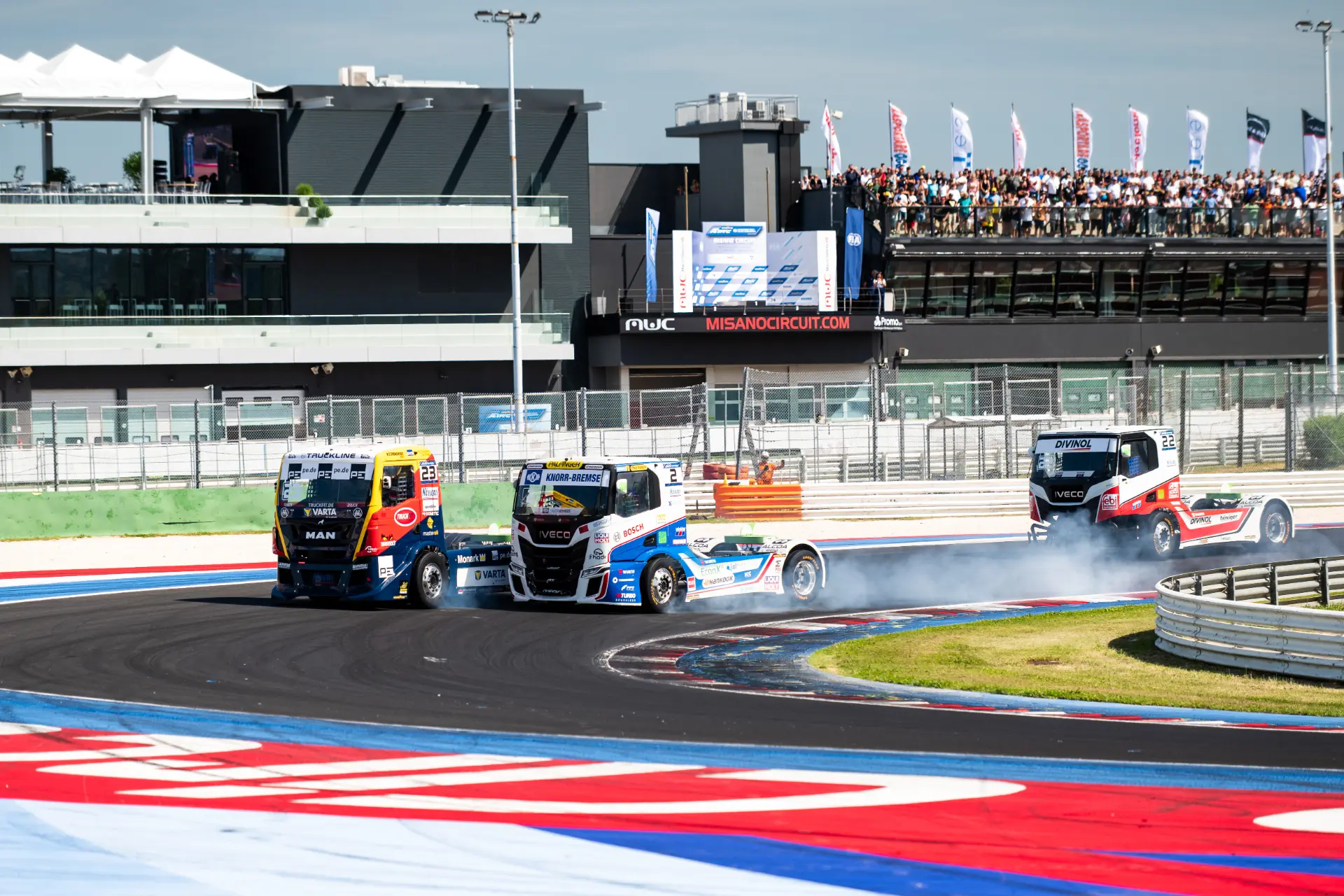 Tres camiones de carreras toman una curva cerrada en la pista de carreras europa misano etrc 2026, echando humo mientras los espectadores observan desde las gradas durante estas emocionantes carreras de camiones.