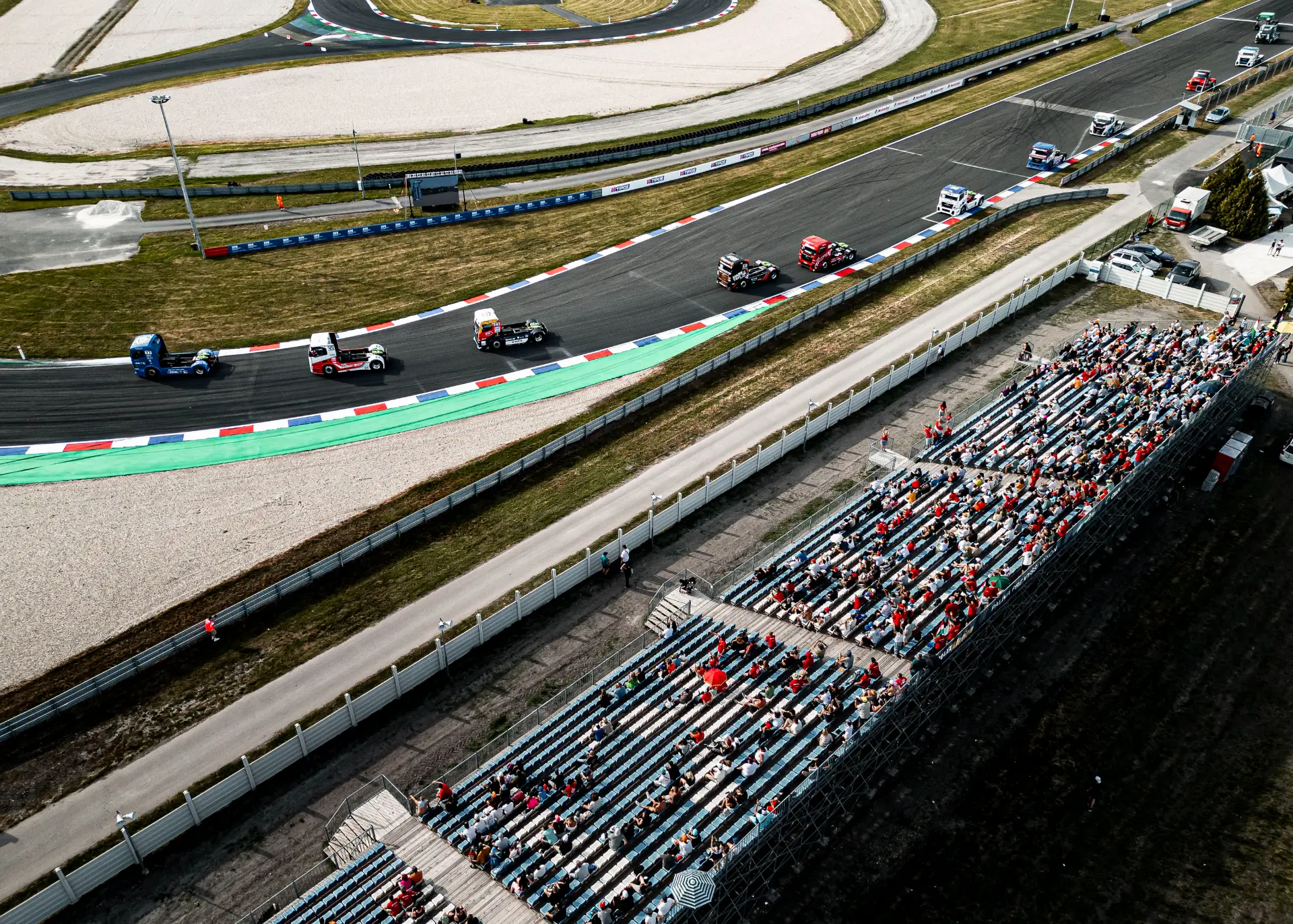 Vista aérea de un circuito de carreras con grandes camiones compitiendo y espectadores en gradas junto a la pista, captada por un sistema Borrador automático.