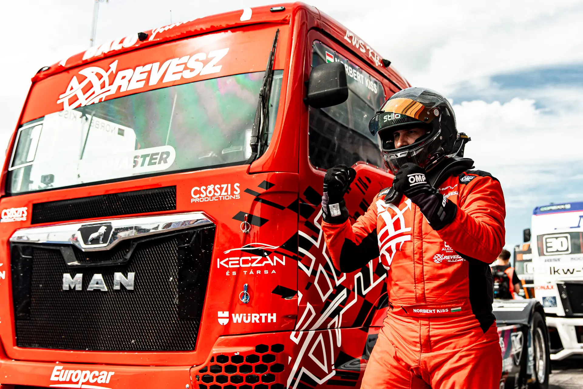 Un piloto de carreras vestido de rojo celebra junto a un camión MAN rojo, mostrando el Borrador automático en un emocionante evento de carreras.