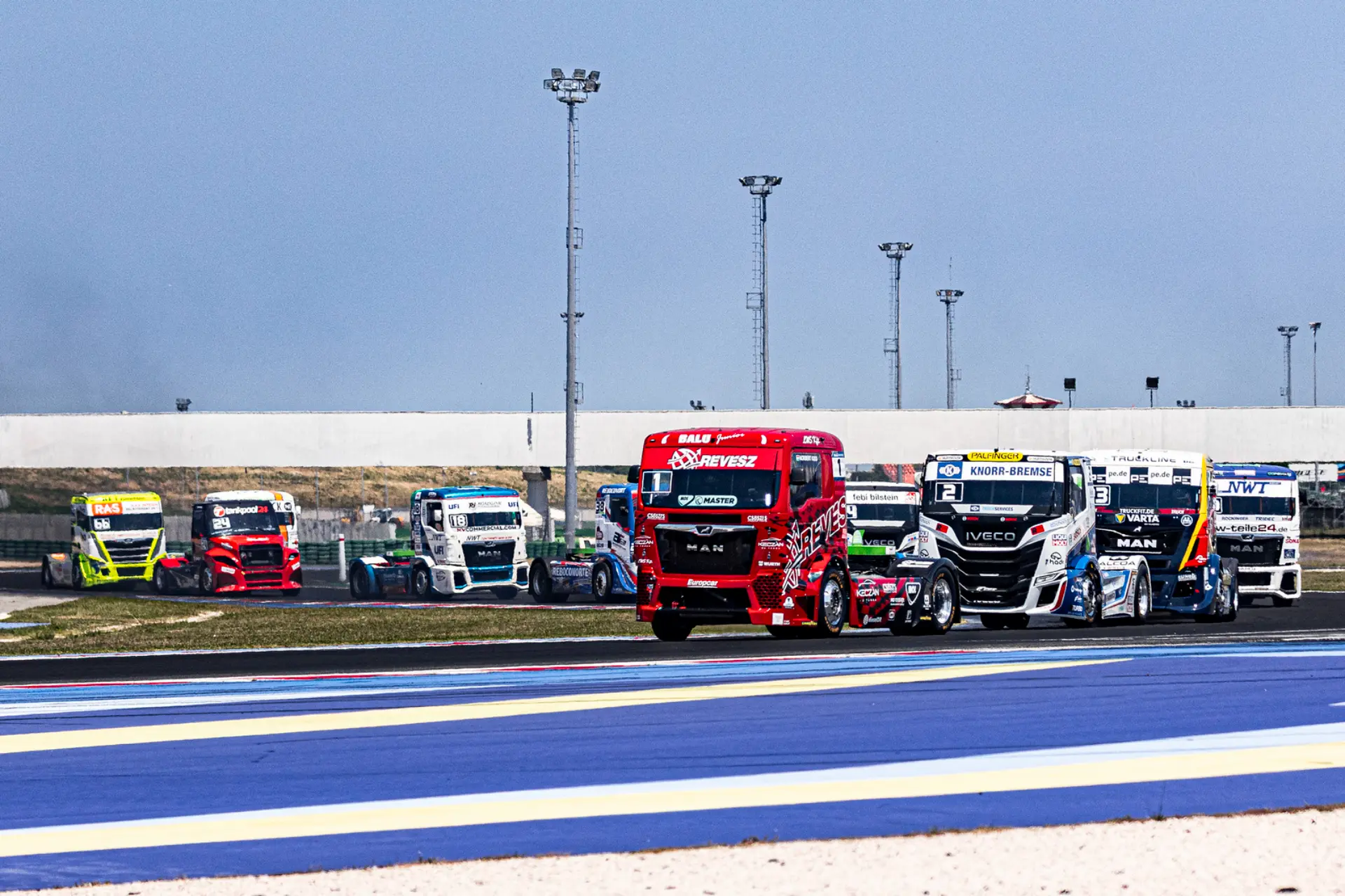 Varios camiones de carreras de colores compiten en una pista de carreras durante el emocionante campeonato europeo carreras camiones 2026, a toda velocidad bajo un puente en un día soleado en Misano ETRC 2026.