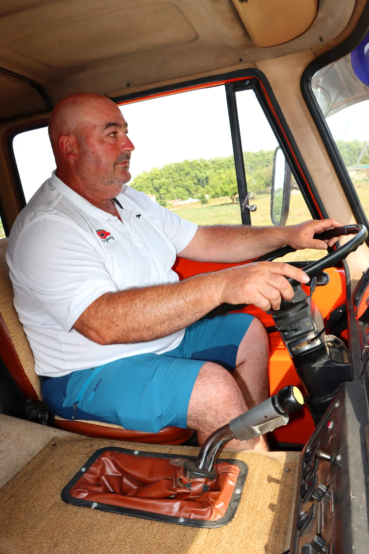 Un hombre con camisa blanca y pantalones cortos azules conduce un vehículo Barreiros 42.20, sujetando el volante y mirando hacia delante.
