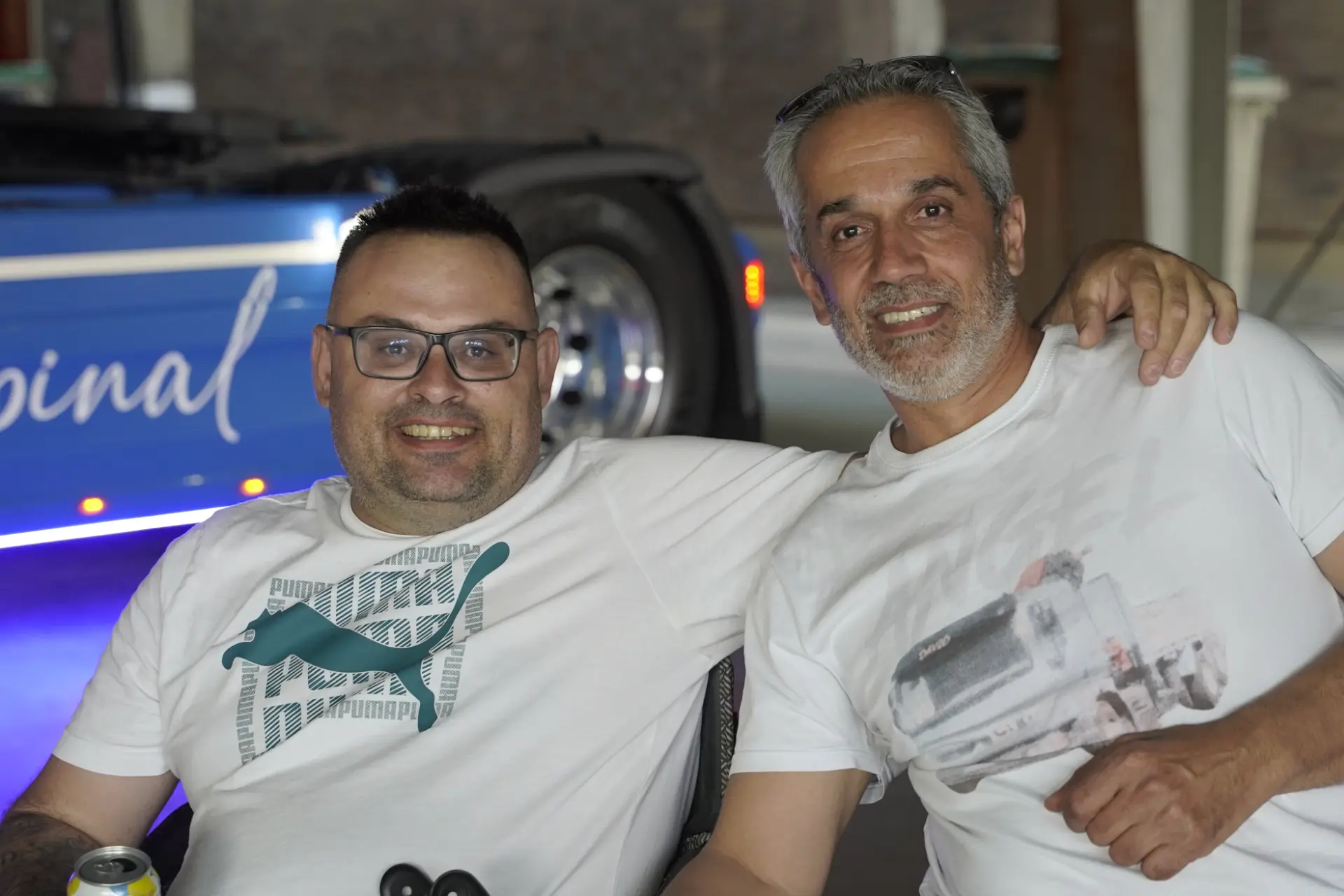 Dos hombres sonrientes con camisetas blancas se sientan juntos, uno con el brazo alrededor del otro, posando para una foto durante la concentración solidaria de camiones en Ávila, rodeados de vehículos.