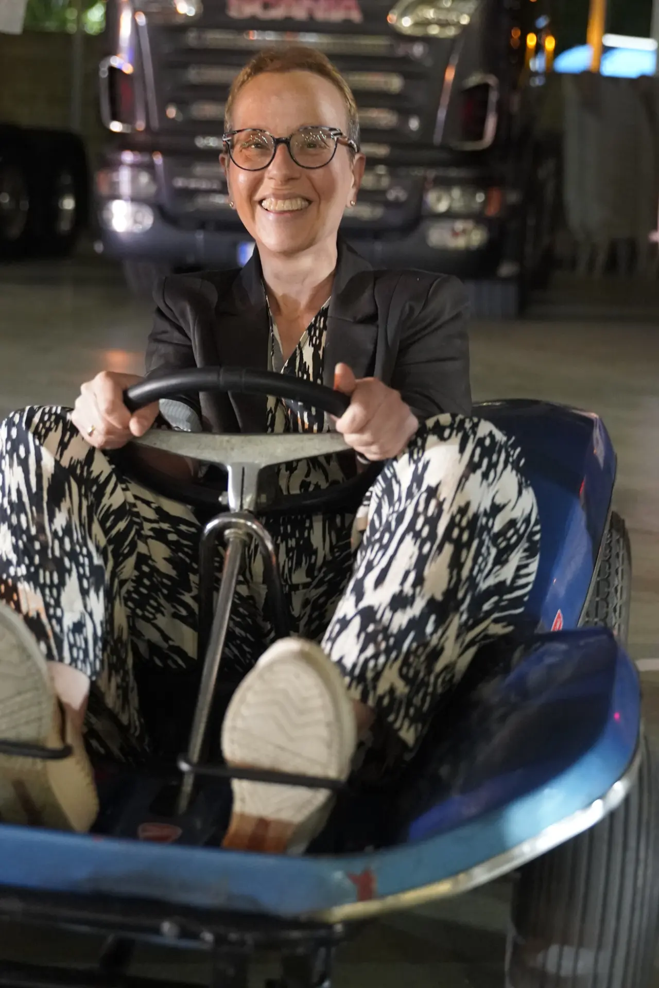 Una persona sonriente con gafas conduce un kart en el interior, con un traje estampado y zapatillas de deporte, mientras los camiones se reúnen cerca para la concentración solidaria de camiones en Ávila.