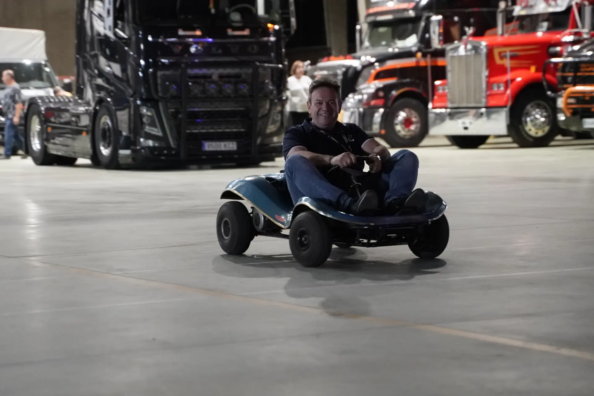 Un hombre conduce un pequeño kart en el interior, sonriente, con grandes camiones de una concentración solidaria de camiones aparcados al fondo.