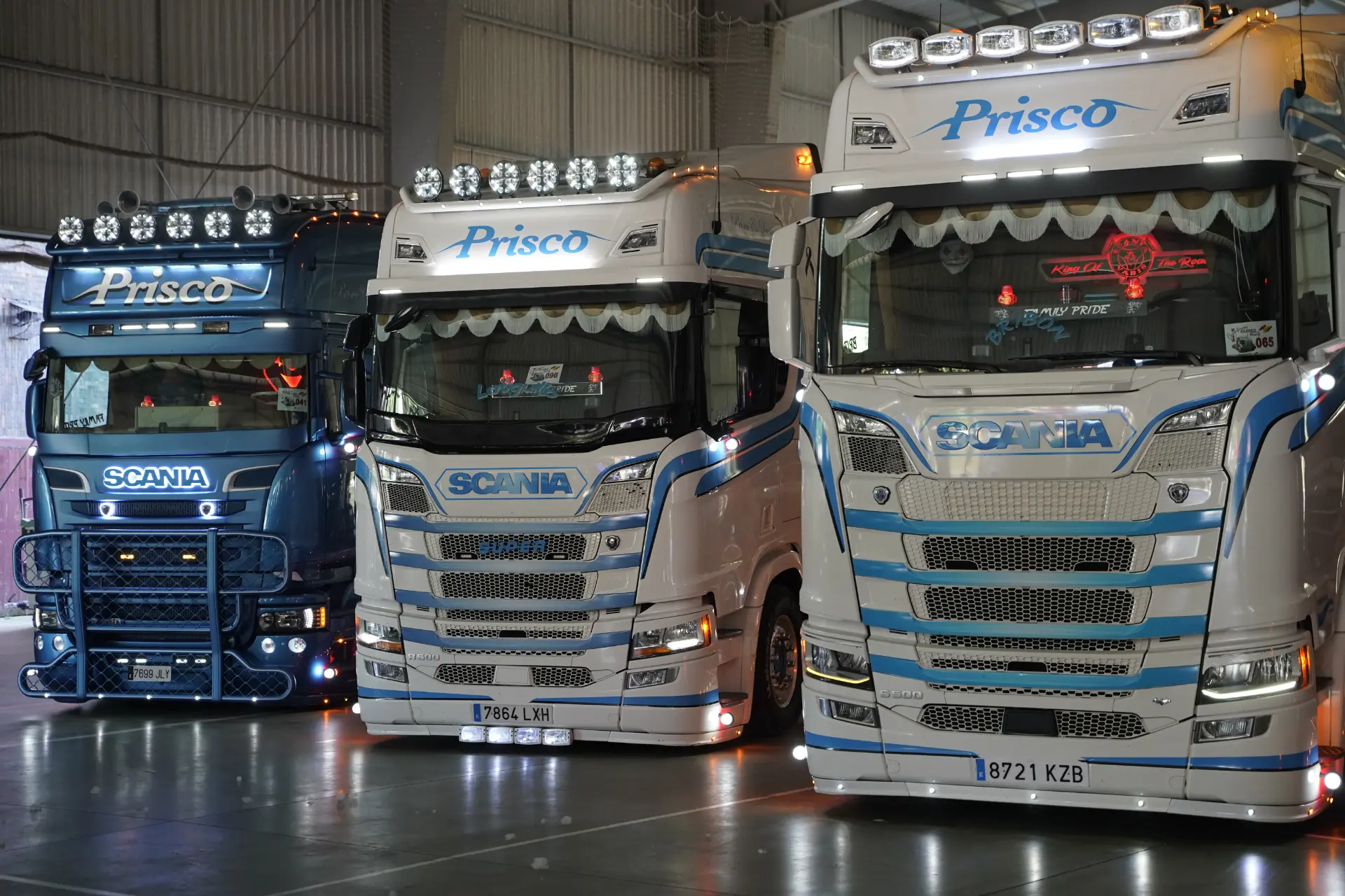Tres vehículos Scania decorados con motivos azules y blancos están aparcados en el interior bajo luces brillantes, listos para la concentración solidaria de camiones en Ávila.