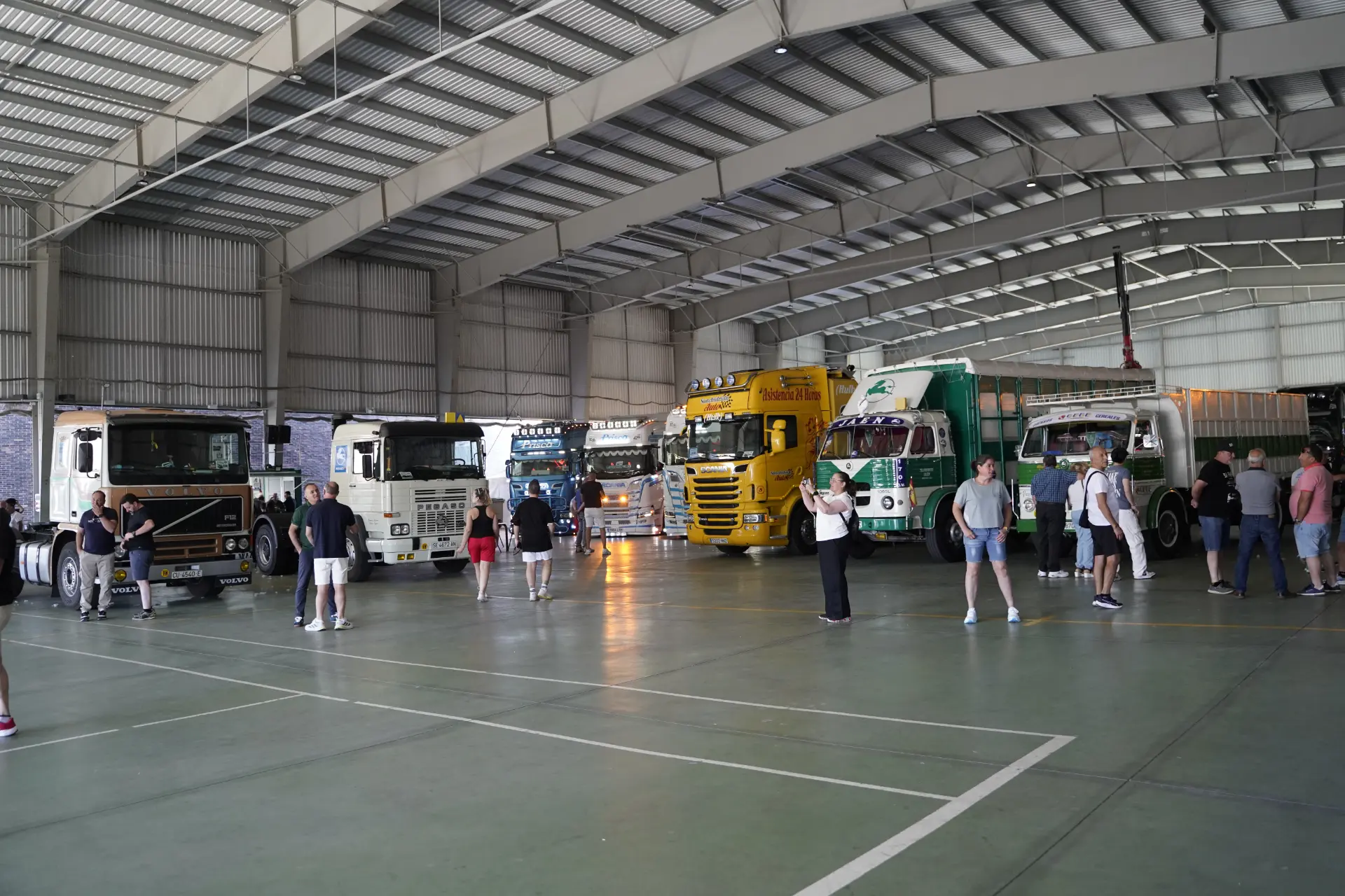 Un grupo de personas permanecen junto a varios vehículos de colores aparcados en el interior de una gran nave industrial durante una concentración solidaria de camiones en Ávila.