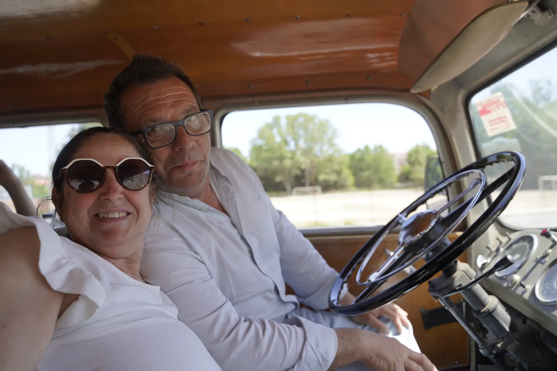 Una mujer y un hombre sonrientes se sientan juntos en el interior de un coche antiguo en un día soleado en la concentración solidaria de camiones de Ávila.