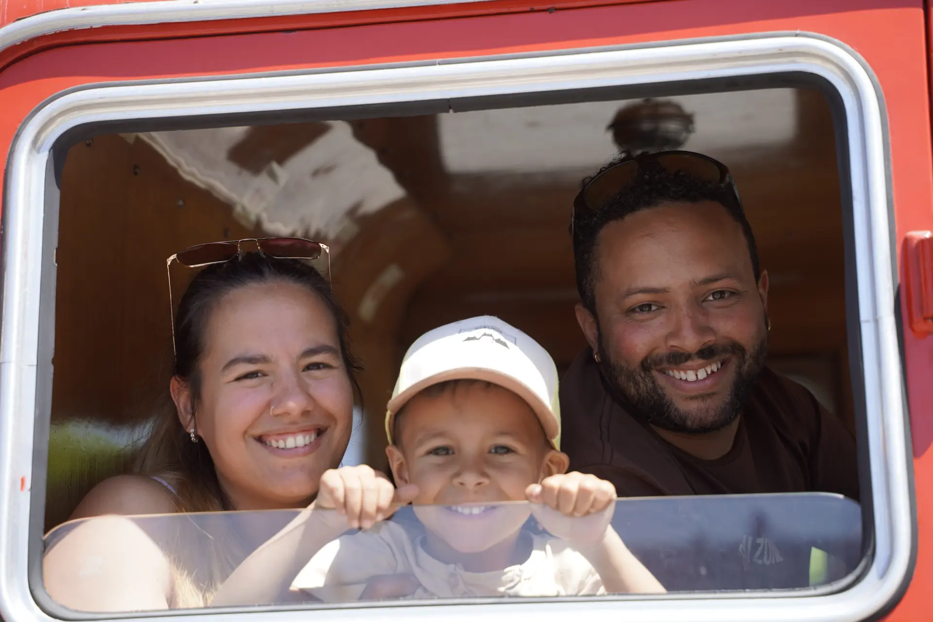 Una sonriente familia de tres miembros mira por la ventanilla de un autobús durante la concentración solidaria de camiones en Ávila; el niño delante, los adultos detrás, todos parecen felices y alegres.