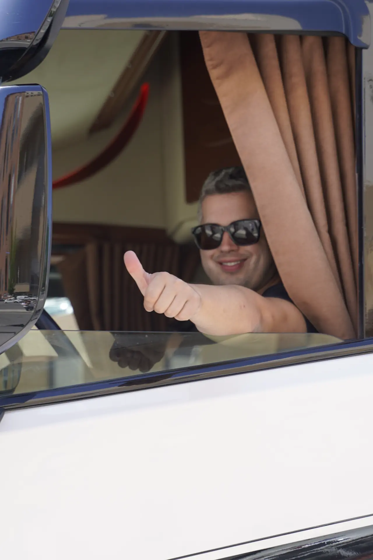 Una persona sonriente con gafas de sol levanta el pulgar sentada en el asiento del conductor de un vehículo durante la concentración solidaria de camiones en Ávila.