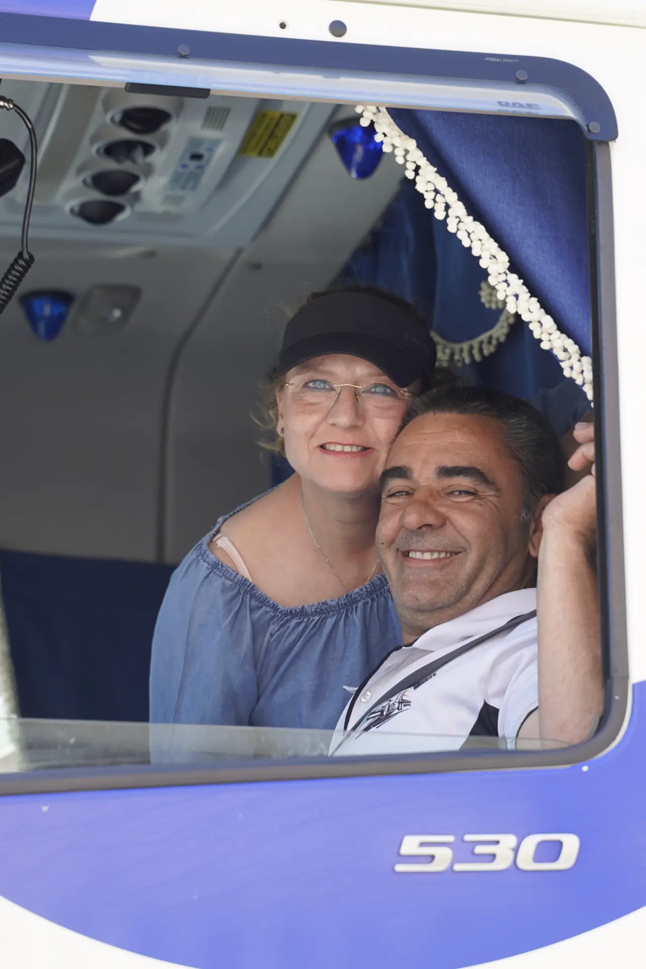 Un hombre y una mujer sonrientes posan juntos en el interior de un vehículo azul y blanco en la concentración solidaria de camiones de Ávila, mirando por una ventana abierta.