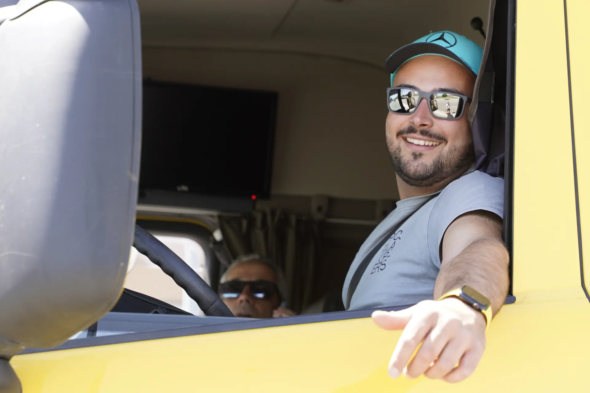 Un hombre sonriente con gafas de sol conduce un camión amarillo en la concentración solidaria de camiones de Ávila, con otra persona sentada en el asiento del copiloto entre otros muchos vehículos.