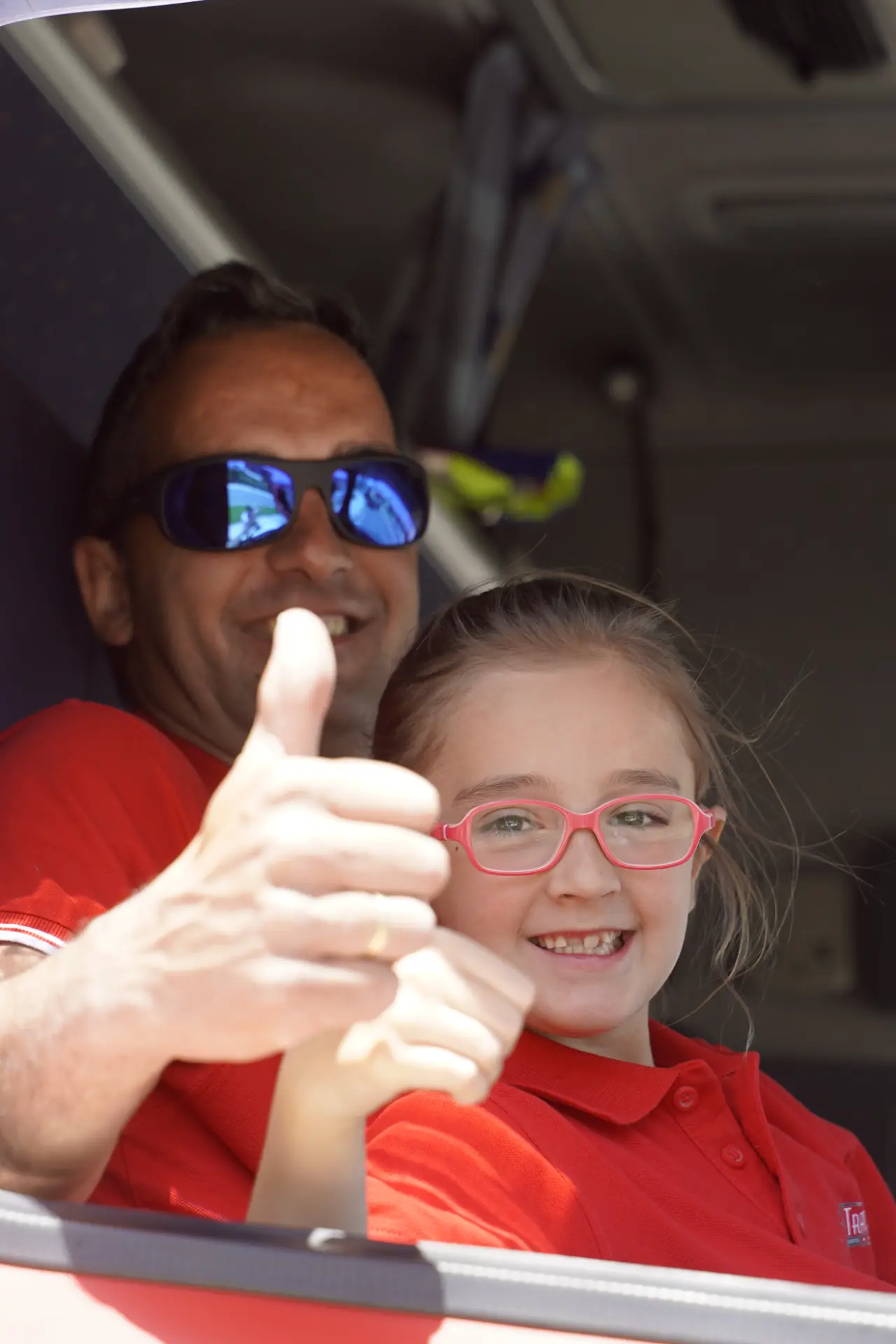 Un hombre y una joven con camiseta roja y gafas sonríen y se saludan con el pulgar mientras están sentados juntos en la concentración solidaria de Ávila, con camiones al fondo.
